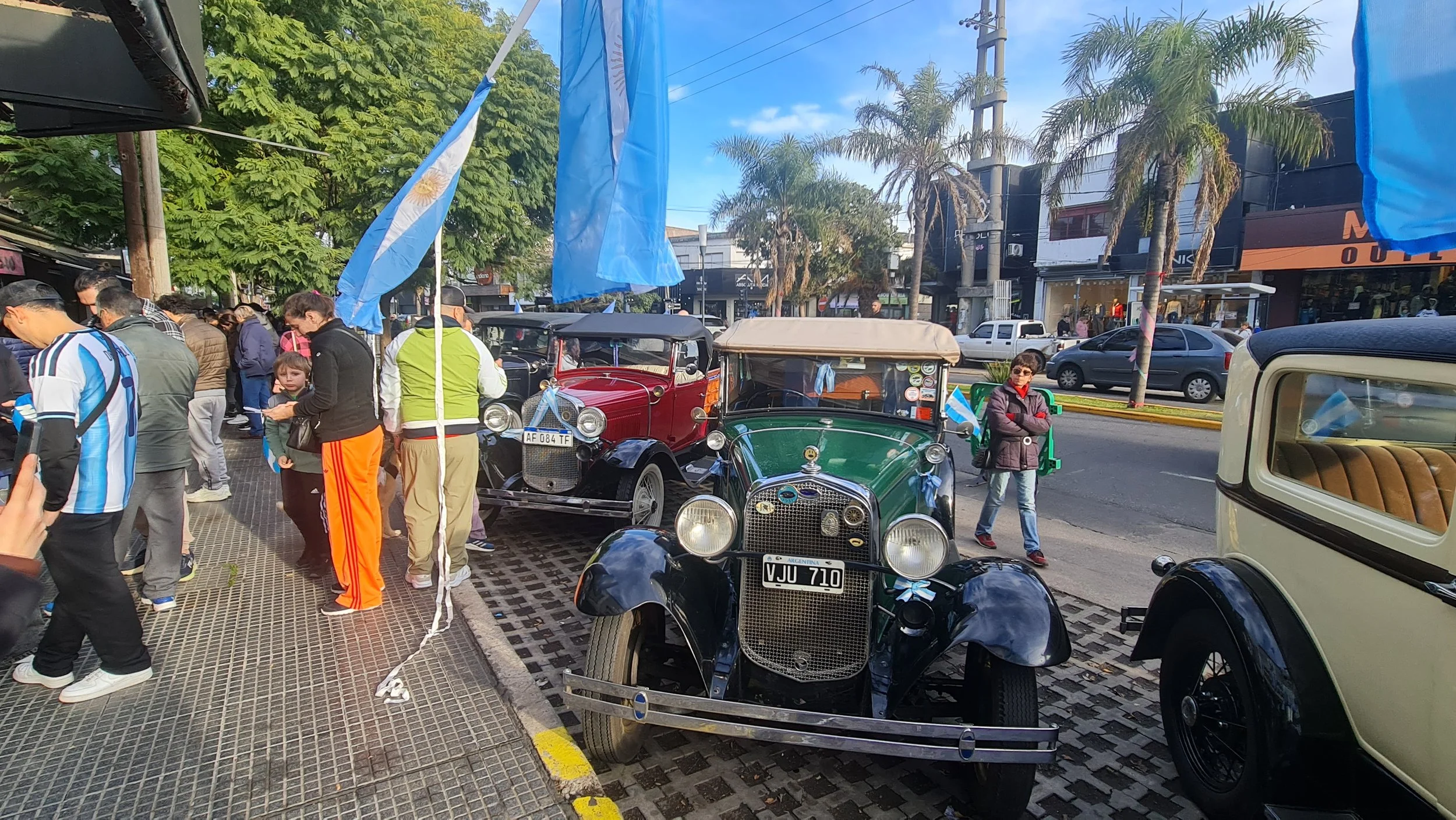 Personas en una calle observando coches clásicos y antiguos, con banderas argentinas colgando de postes, en un día soleado con árboles y edificios en el fondo.