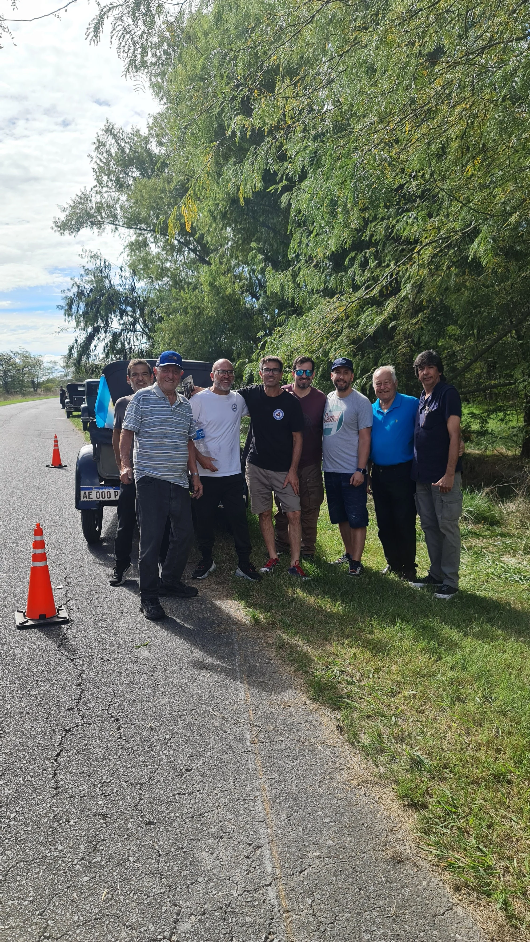 Un grupo de ocho hombres joven, en una calle rural, posando para la foto. Algunos llevan gorras, camiseta y lentes oscuros. Hay conos de señalización en la carretera y un vehículo de fondo. Rodeados de árboles y vegetación.