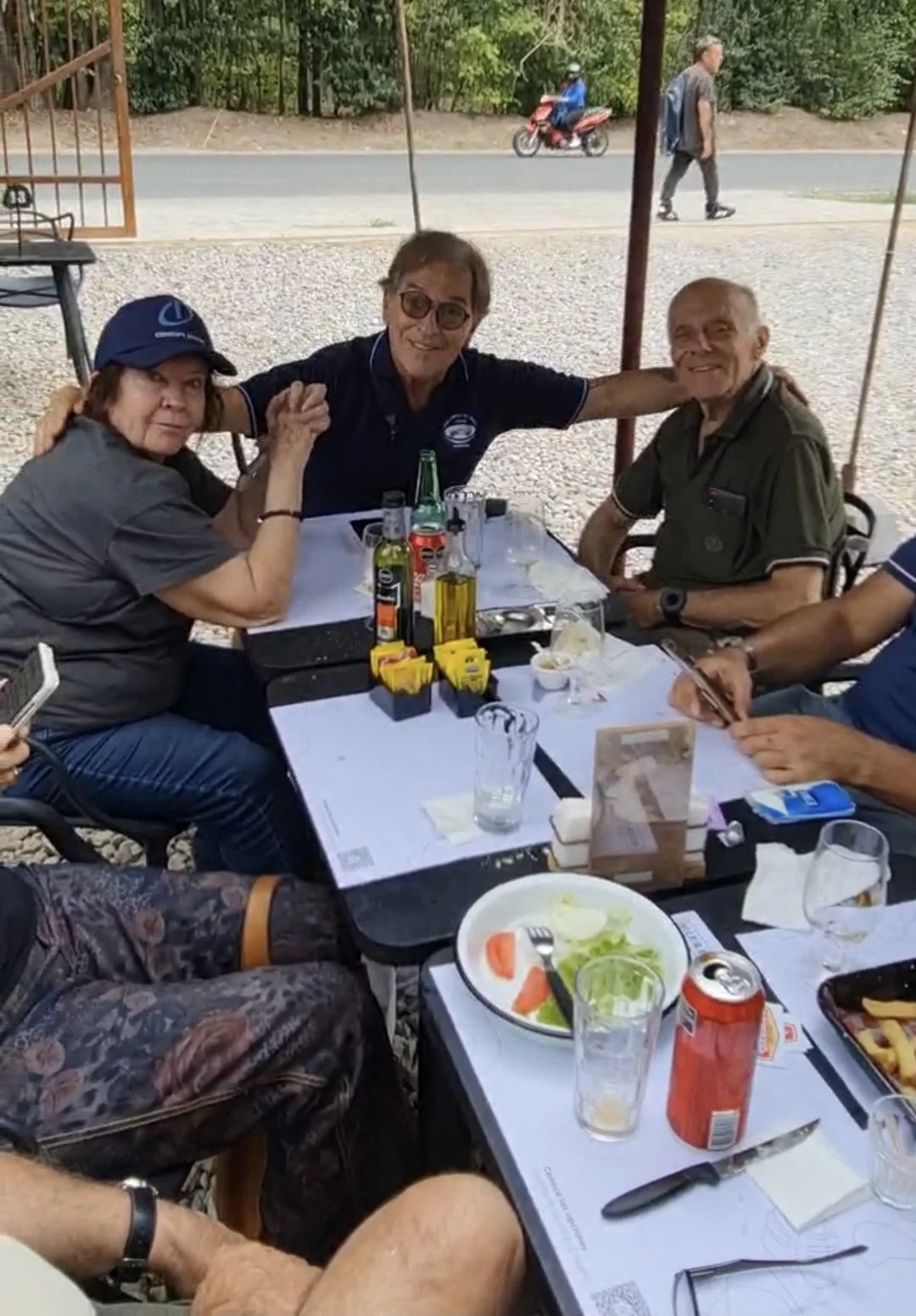 Grupo de personas sentadas en una mesa al aire libre, disfrutando de comida y bebida, con vegetación y una calle en el fondo.