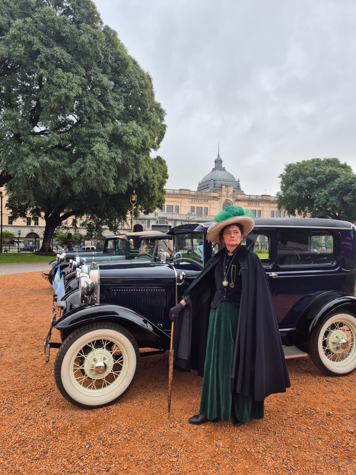 Mujer vestida con ropa antigua y sombrero grande de colores verdes, parada junto a un coche clásico en un parque con árboles y un edificio histórico de fondo.