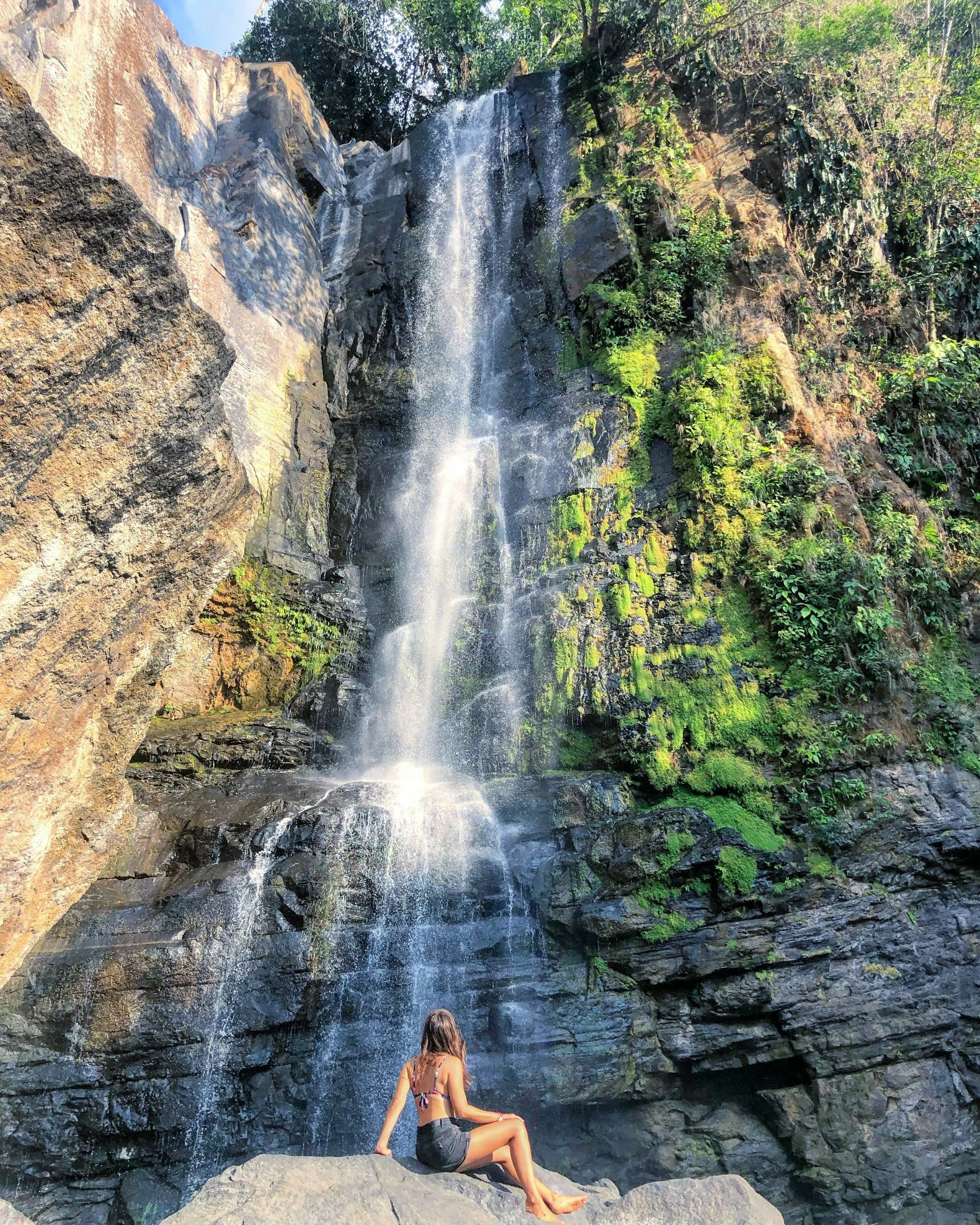 A woman sitting on rocks at the base of a waterfall surrounded by lush greenery.