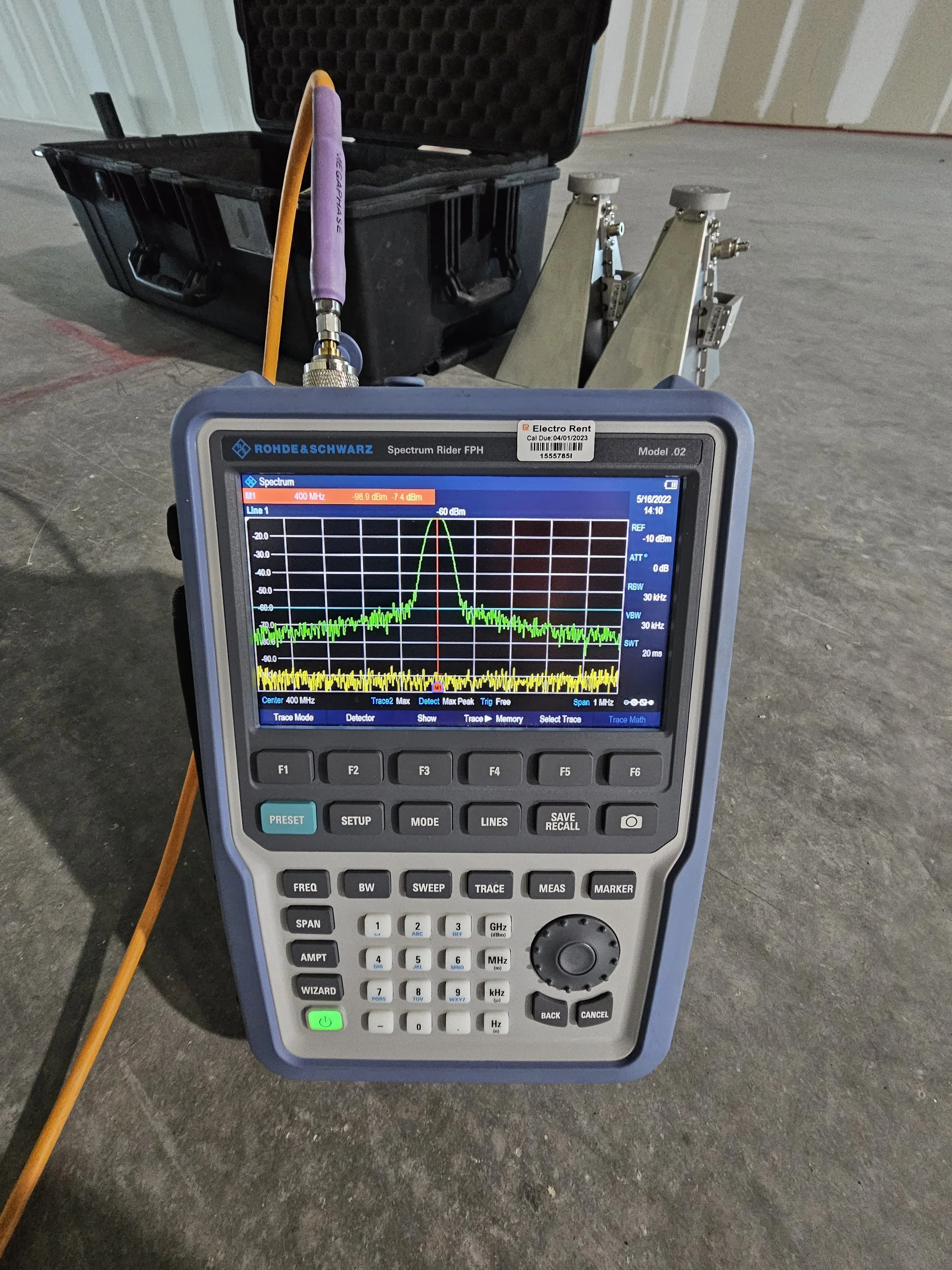 A spectrum analyzer displaying a frequency peak at 400 MHz, connected to a yellow and purple antenna, with metal tripod stands and a black hard case in the background, set up in a construction or industrial site.