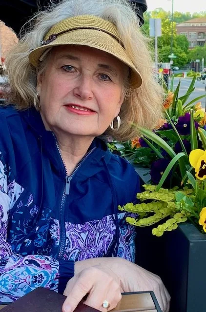 A woman with curly blonde hair wearing a tan visor and a blue jacket, sitting outdoors next to colorful flowers, in what appears to be a garden center or park.