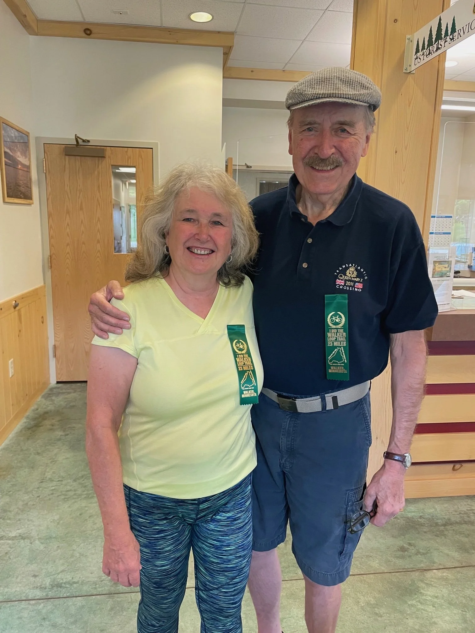 A smiling woman and man standing side by side indoors, both wearing green ribbons that read 'WALKER CROSSING 5K 2011 WALKER, MINNESOTA.' The woman is wearing a light yellow shirt and patterned leggings, and the man is wearing a dark polo shirt, shorts, and a cap.