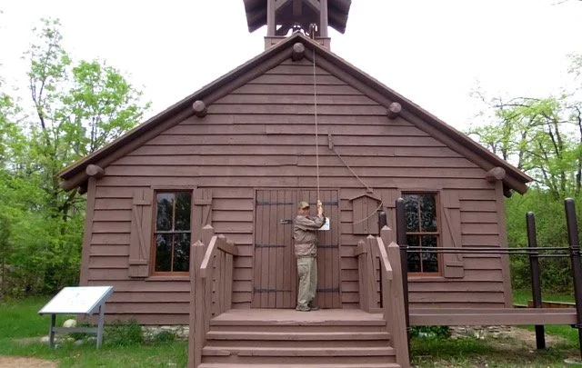 Person hanging a bell on the front of a small wooden church building with steps, two windows, and surrounding trees.