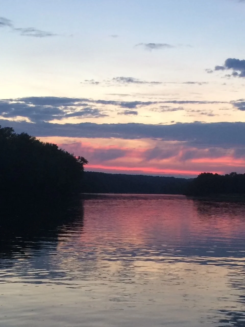 Sunset over a calm river with trees along the banks, sky filled with clouds and pink, purple, and blue hues reflecting on the water.
