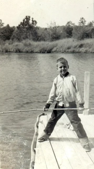 A young boy standing on a small boat or dock, holding a fishing rod, with a river and trees in the background.