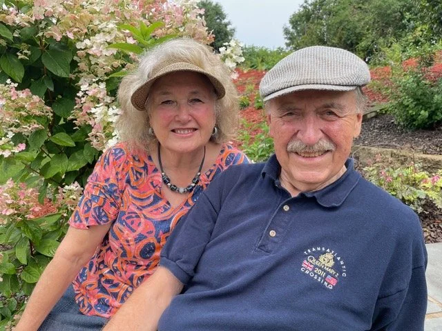 An elderly man and woman smiling outdoors, with blooming pink and white hydrangeas and greenery in the background.
