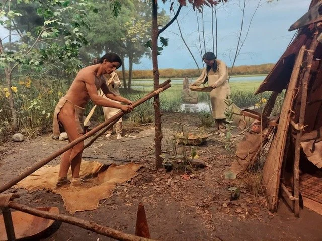 Three people working outdoors near water, with one person digging and the others observing or preparing materials, in a rural setting with trees and makeshift shelter.
