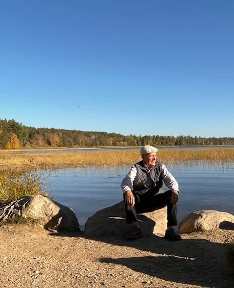 A man sitting on a large rock by a lake, with trees in the background and a clear blue sky.