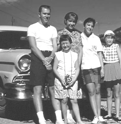 A vintage black and white photo of five children standing in front of a car outdoors.