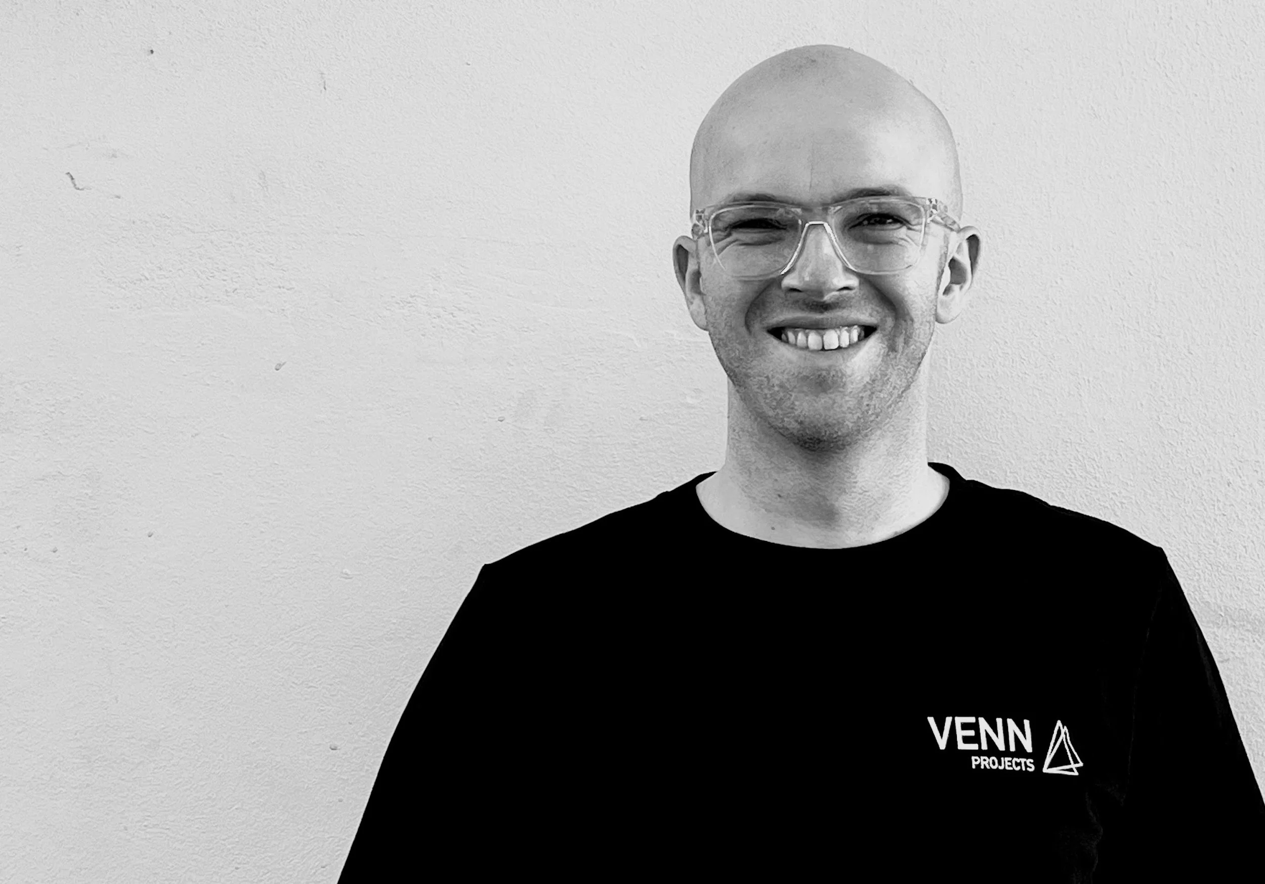 A smiling man with glasses and a shaved head wearing a black shirt with the "VENN PROJECTS" logo stands against a plain white wall.