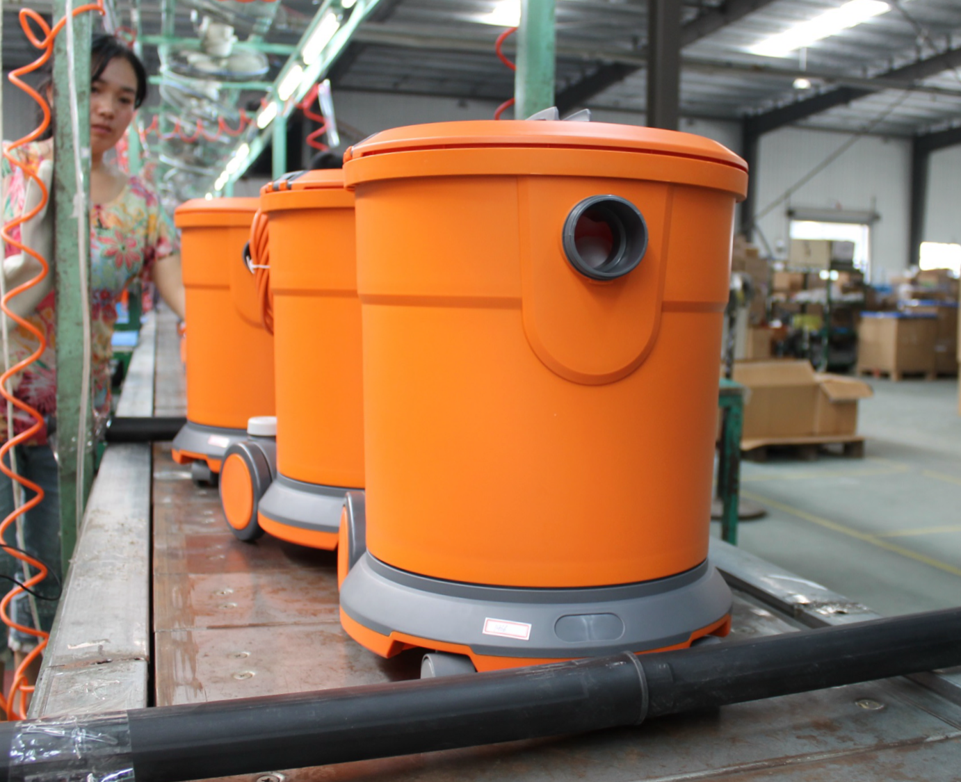 Three orange industrial vacuum cleaners on a production line in a warehouse, with shelves and boxes in the background.