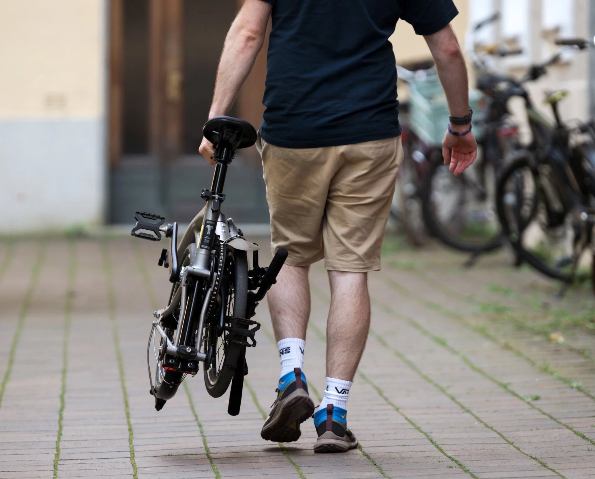 A man walking outdoors carrying a folded bicycle in his right hand, wearing beige shorts, a black T-shirt, and athletic shoes.