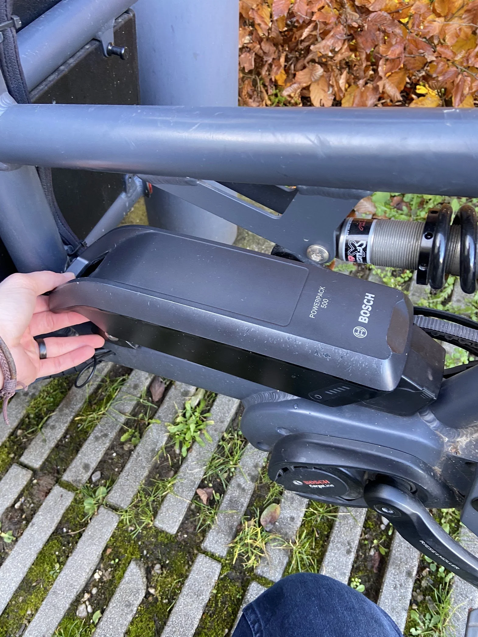 Close-up of an electric bicycle with a Bosch PowerPack 500 battery being inserted or removed, outdoors on a concrete surface with grass and fallen leaves.