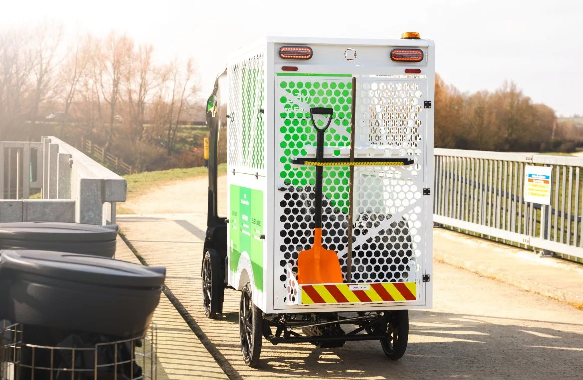 A street-cleaning electric vehicle with a shovel mounted on the back, parked on a bridge with a railing and trees in the background.