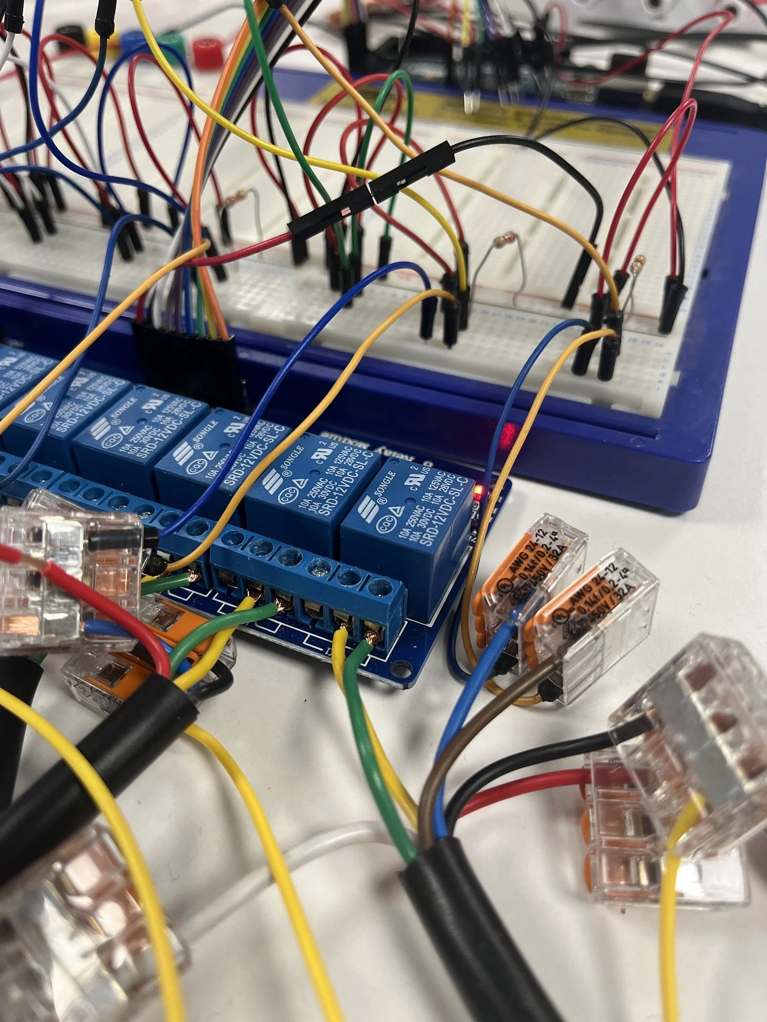 Close-up of an electronic circuit board with multiple colorful wires, relays, and a breadboard connected to a blue device, likely a relay module, on a white surface.