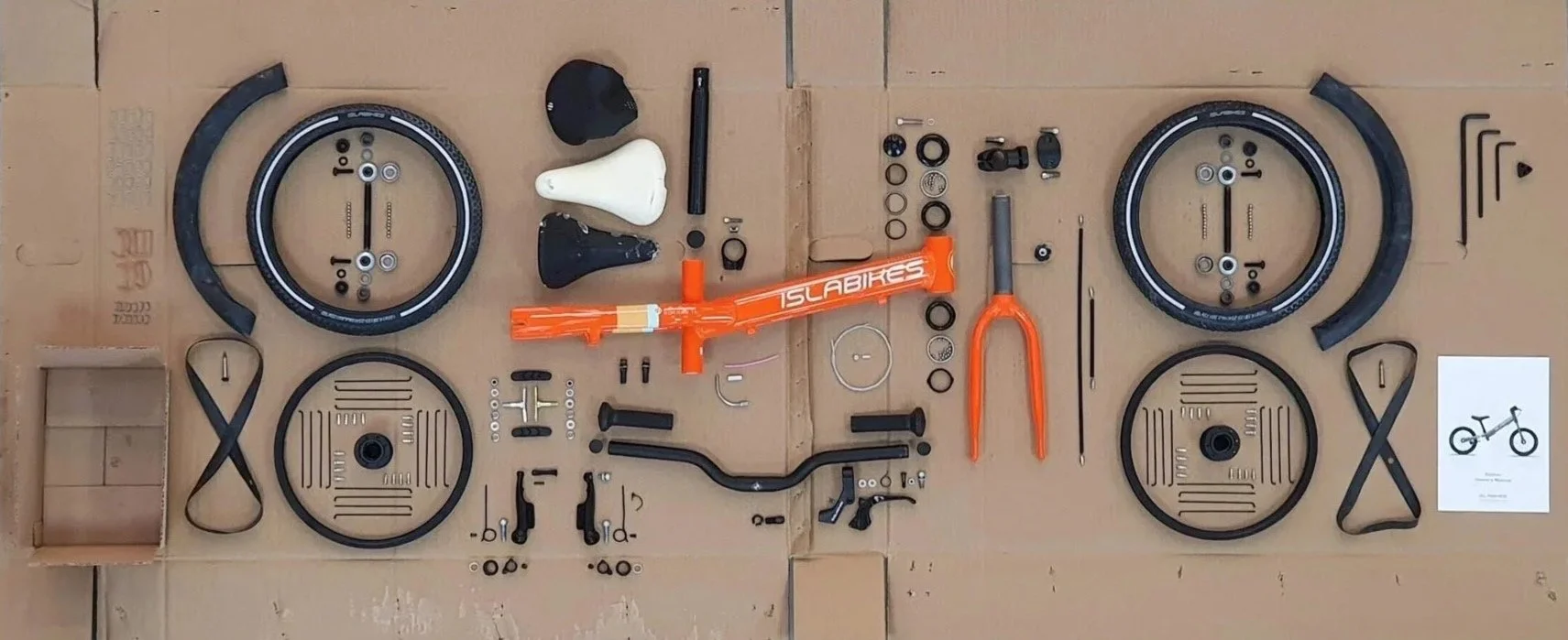 Disassembled orange bicycle frame and various bicycle parts arranged on a cardboard surface, including wheels, pedals, handlebars, and small components.