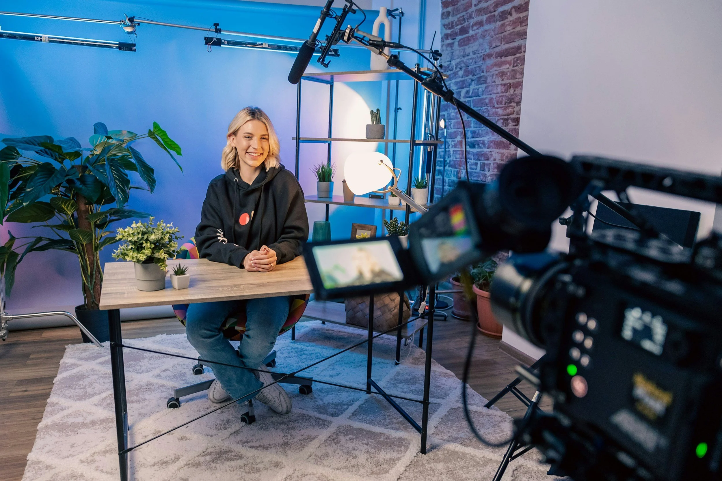 A woman in a black hoodie sits at a wooden desk with plants, surrounded by filming equipment in a cozy, modern studio with blue and purple lighting.