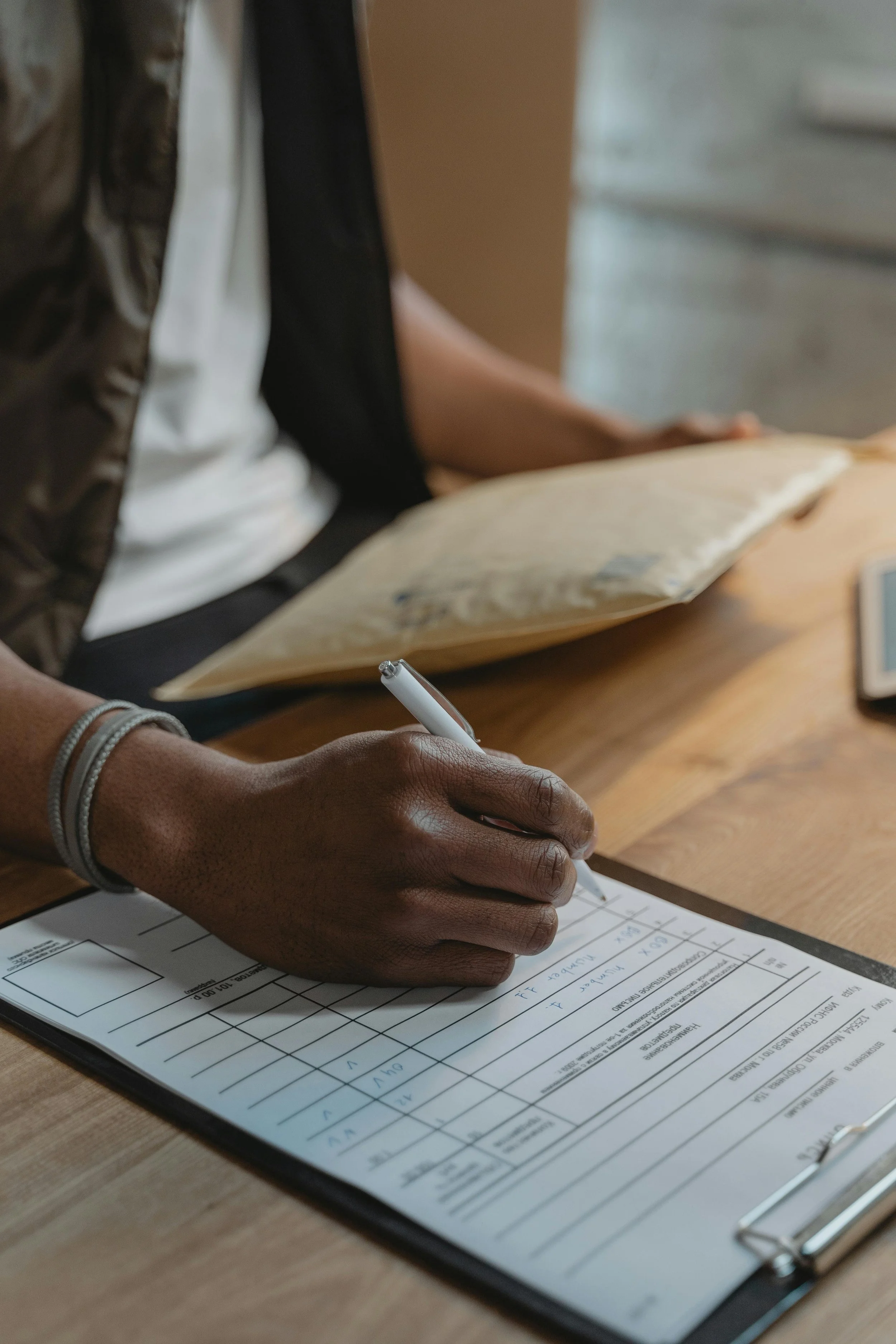 A person writing on a clipboard with a pen, with a paper and smartphone nearby on a wooden table.