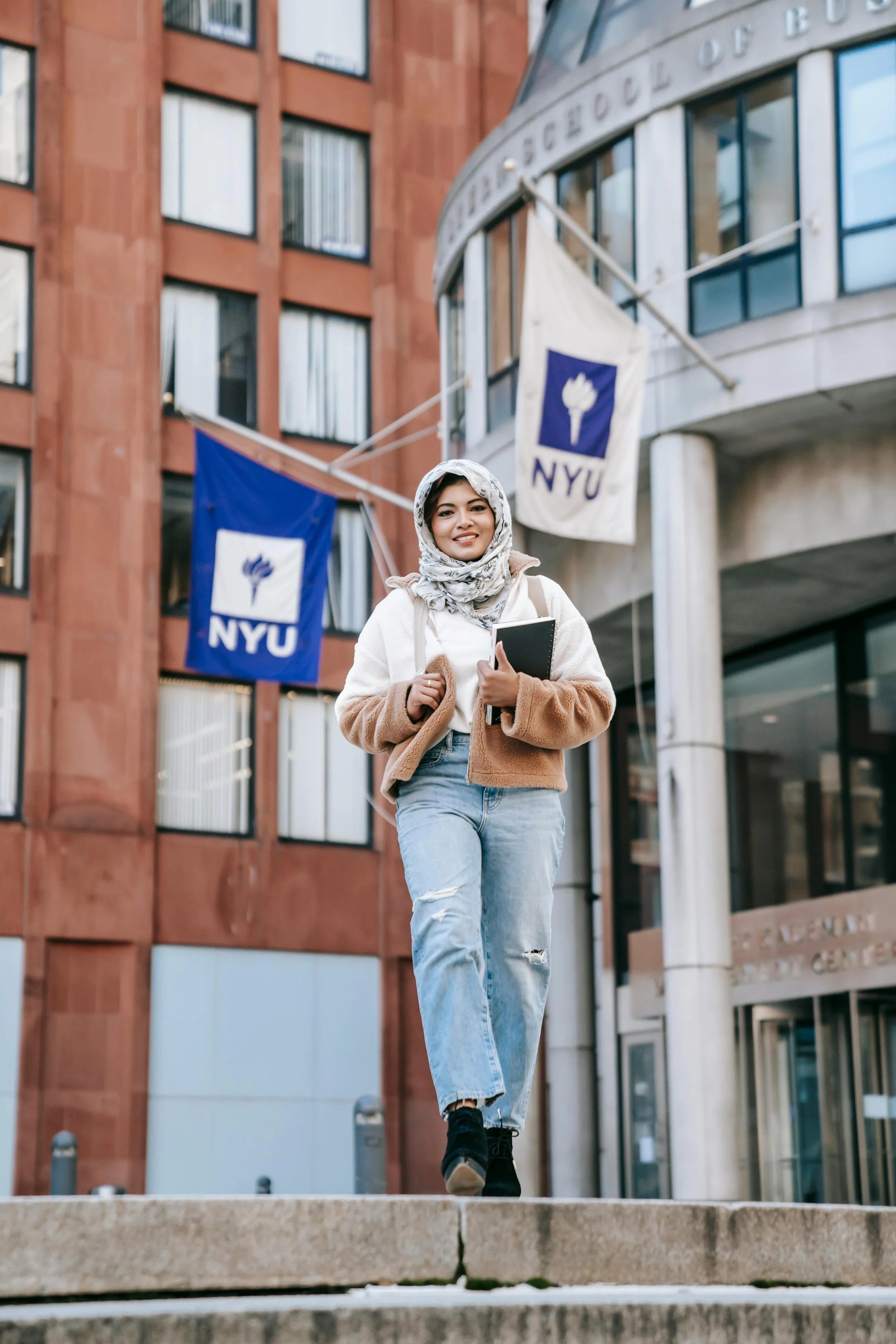 A smiling young woman wearing a headscarf and casual clothes, walking down steps outside NYU building with flags in the background.
