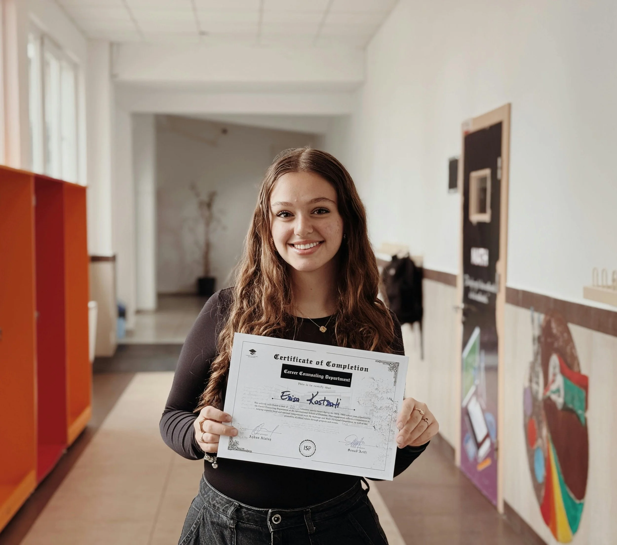 A young woman with long, curly brown hair holding a certificate of completion and smiling inside a school hallway.