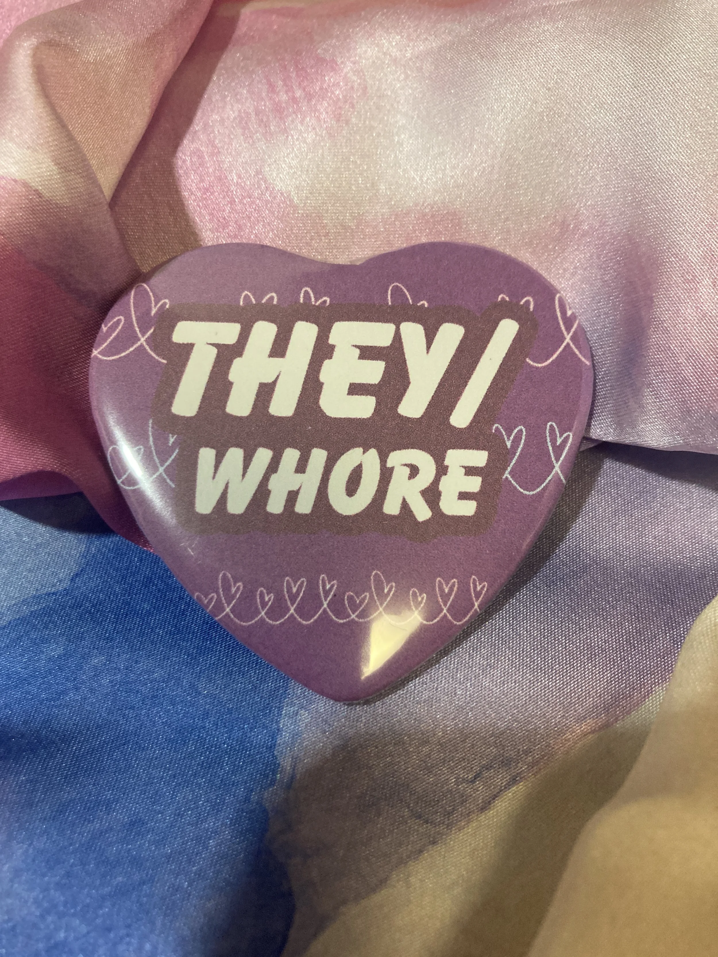 Heart shaped button that says They/Whore with a purple and heart pattern background