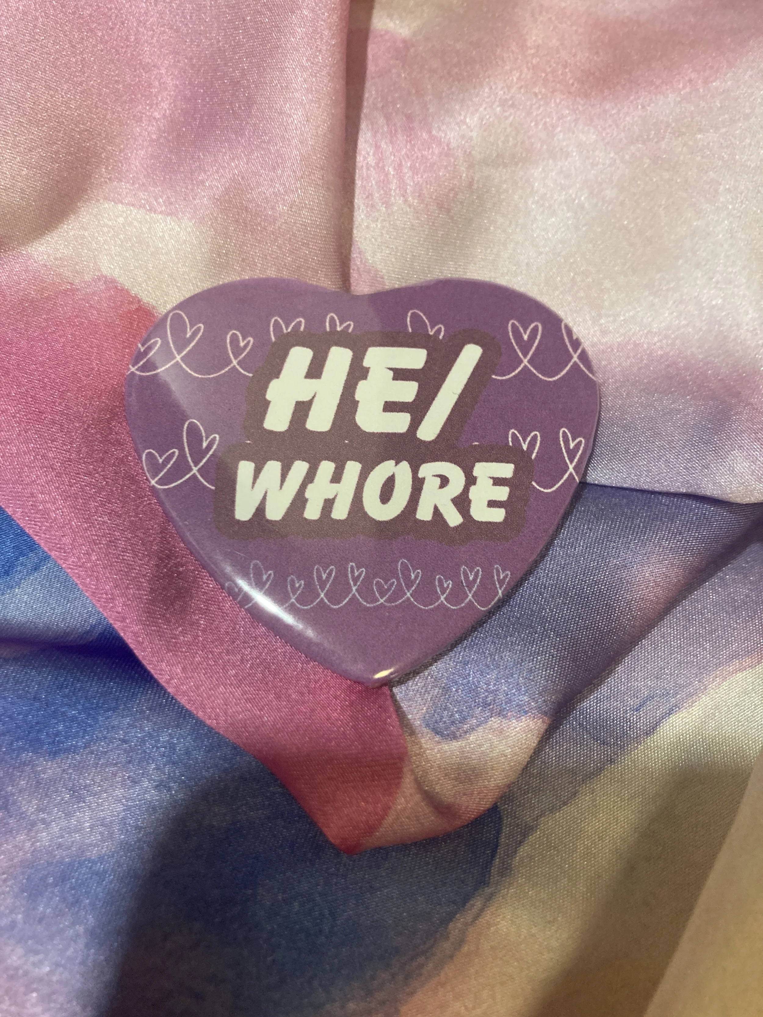 Heart shaped button that says He/Whore with a purple and heart pattern background