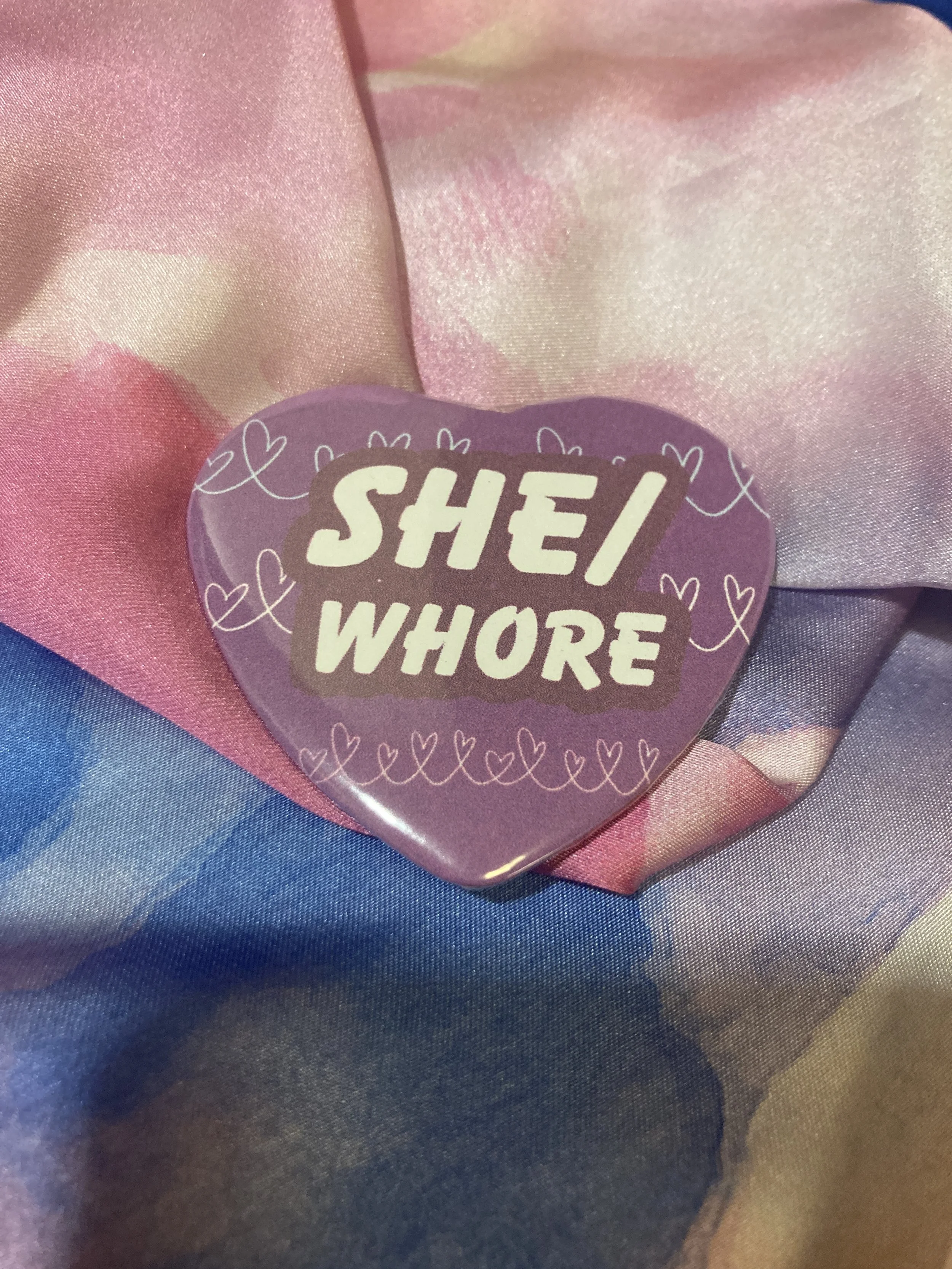Heart shaped button that says She/Whore with a purple and heart pattern background