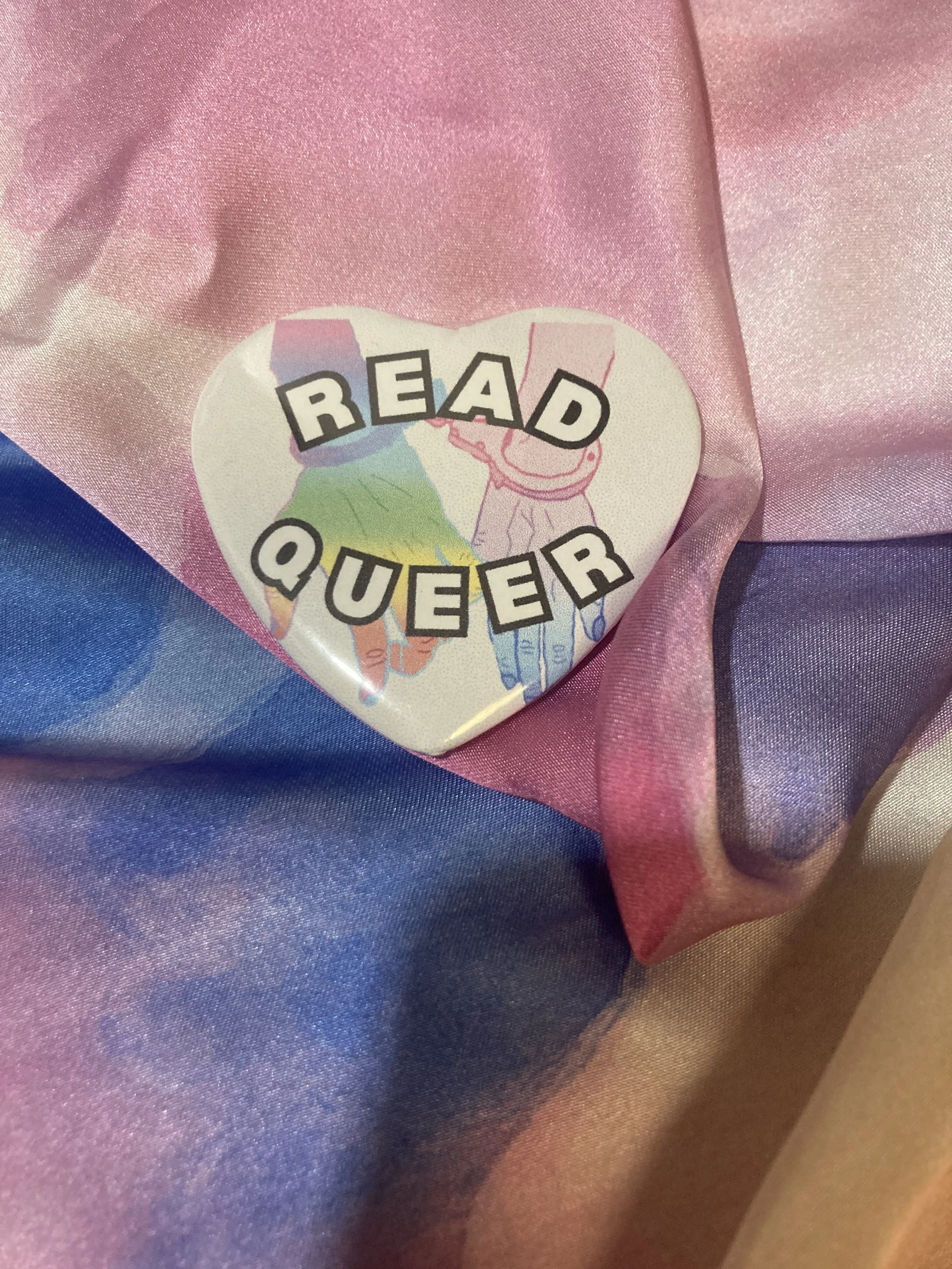 Heart shaped button that says Read Queer with two colorful hands connected by cuffs in the background