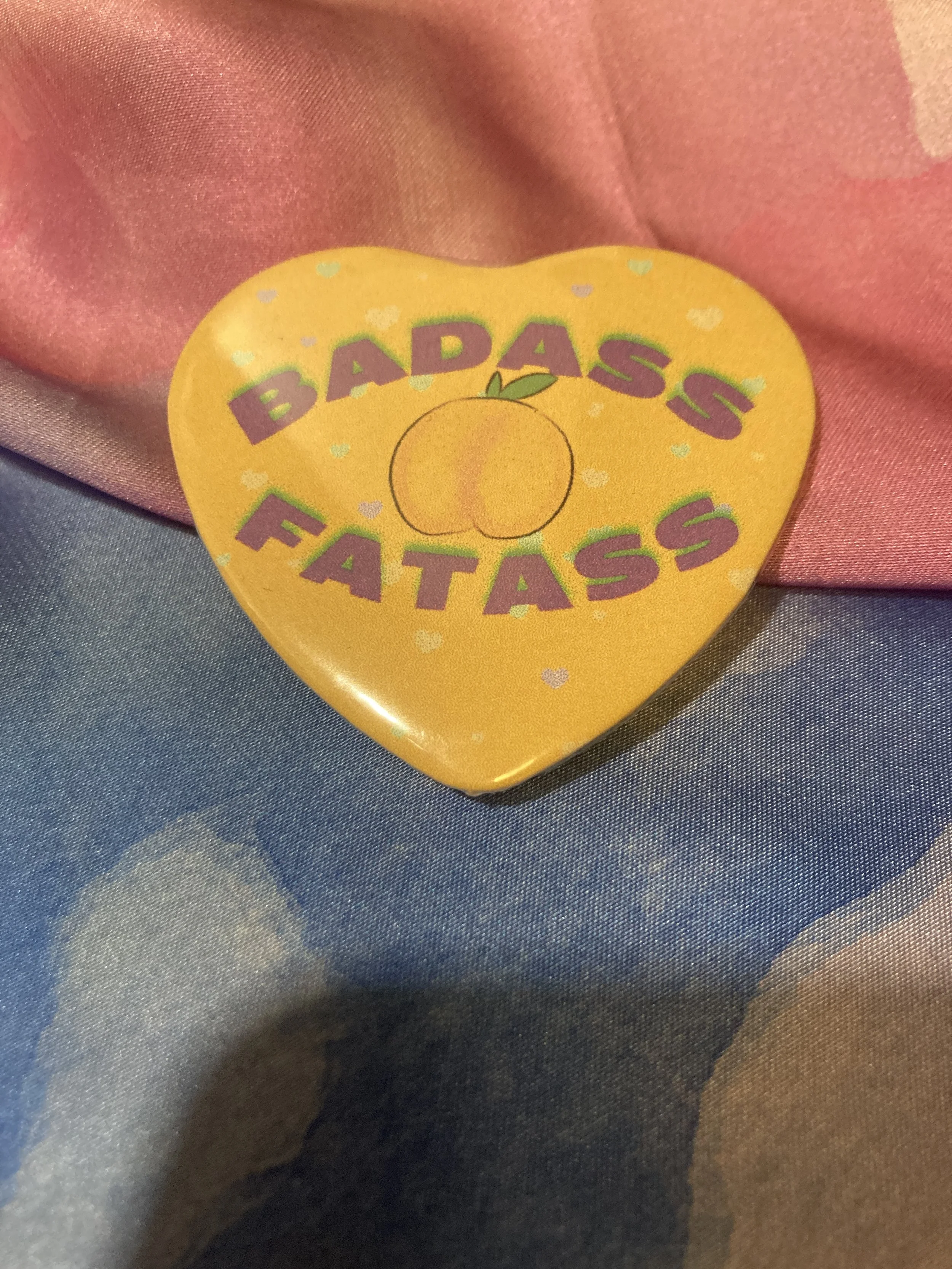 Heart shaped button that says Badass Fatass with an image of a peach on an orange background