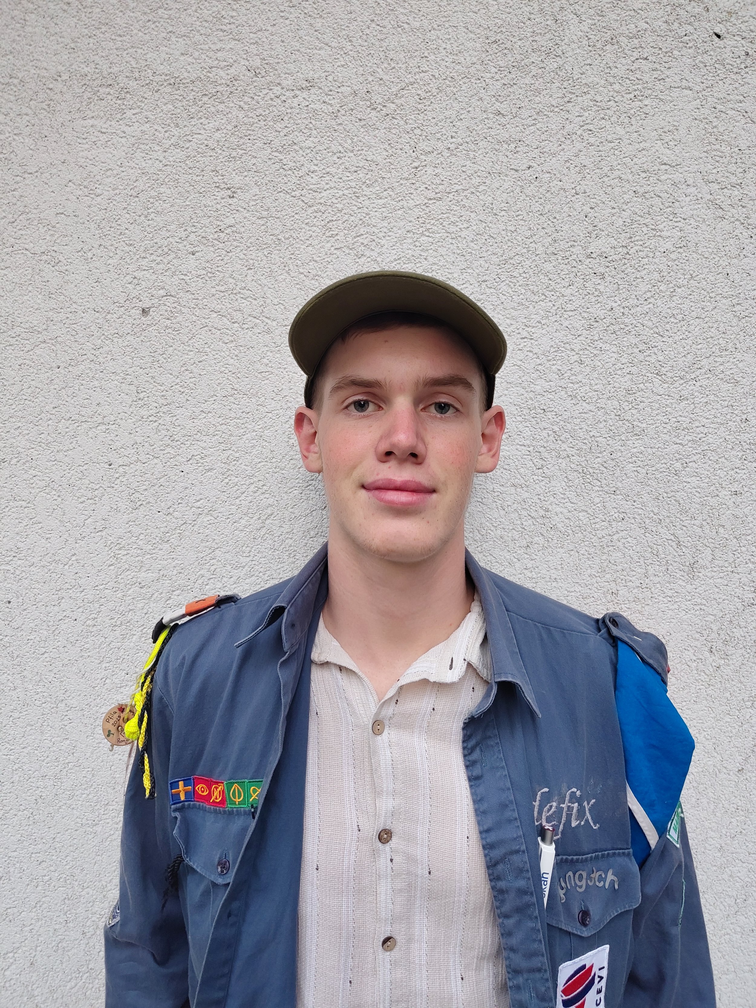 Ein junger Mann trägt eine Mütze und eine Uniform, lächelt leicht vor einem hellen Hintergrund.