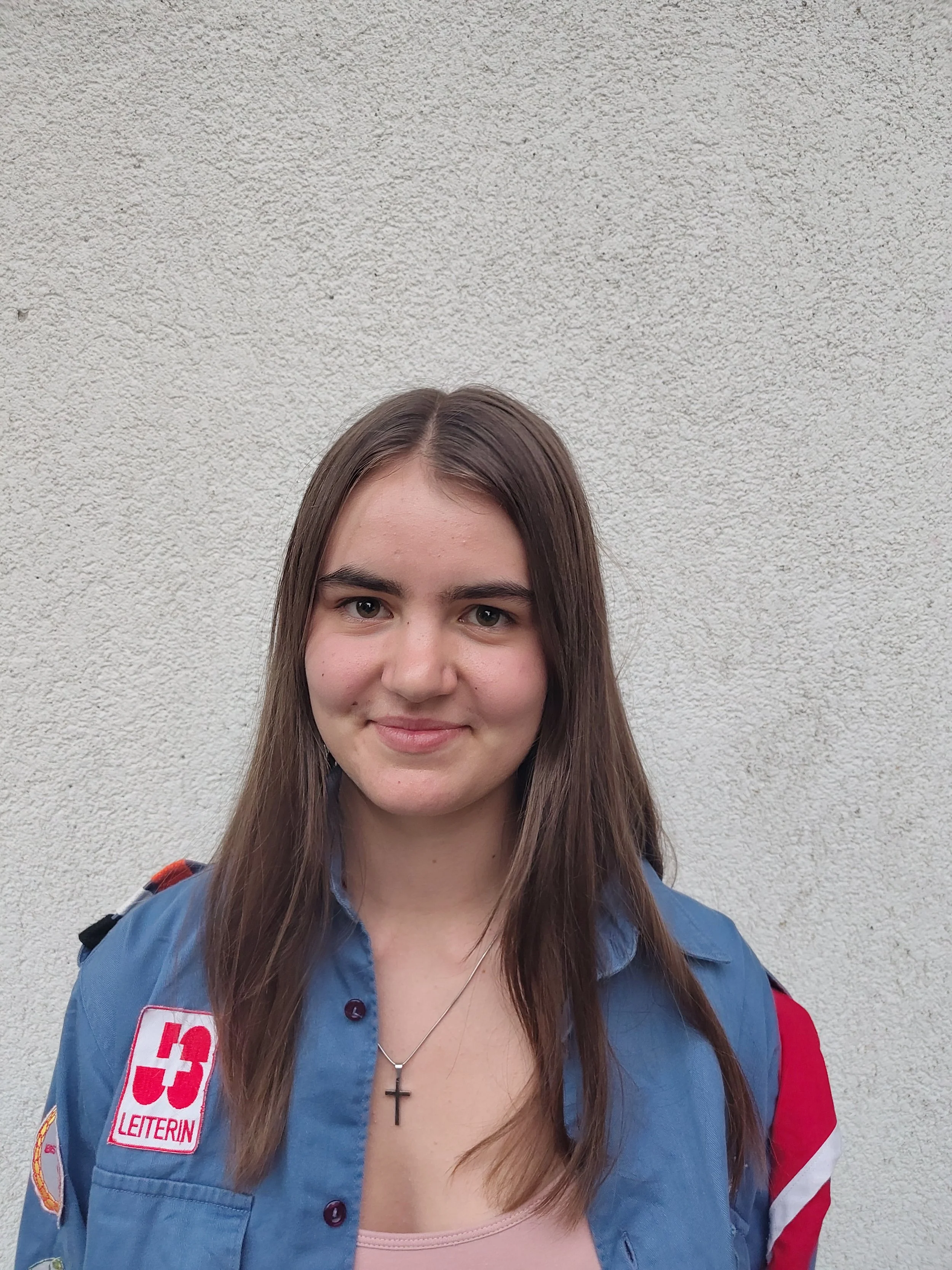 Jugendliches Mädchen mit braunen Haaren in einem Haargummi, trägt eine grüne T-Shirt und eine graue Jacke, steht vor einer weißen Wand.