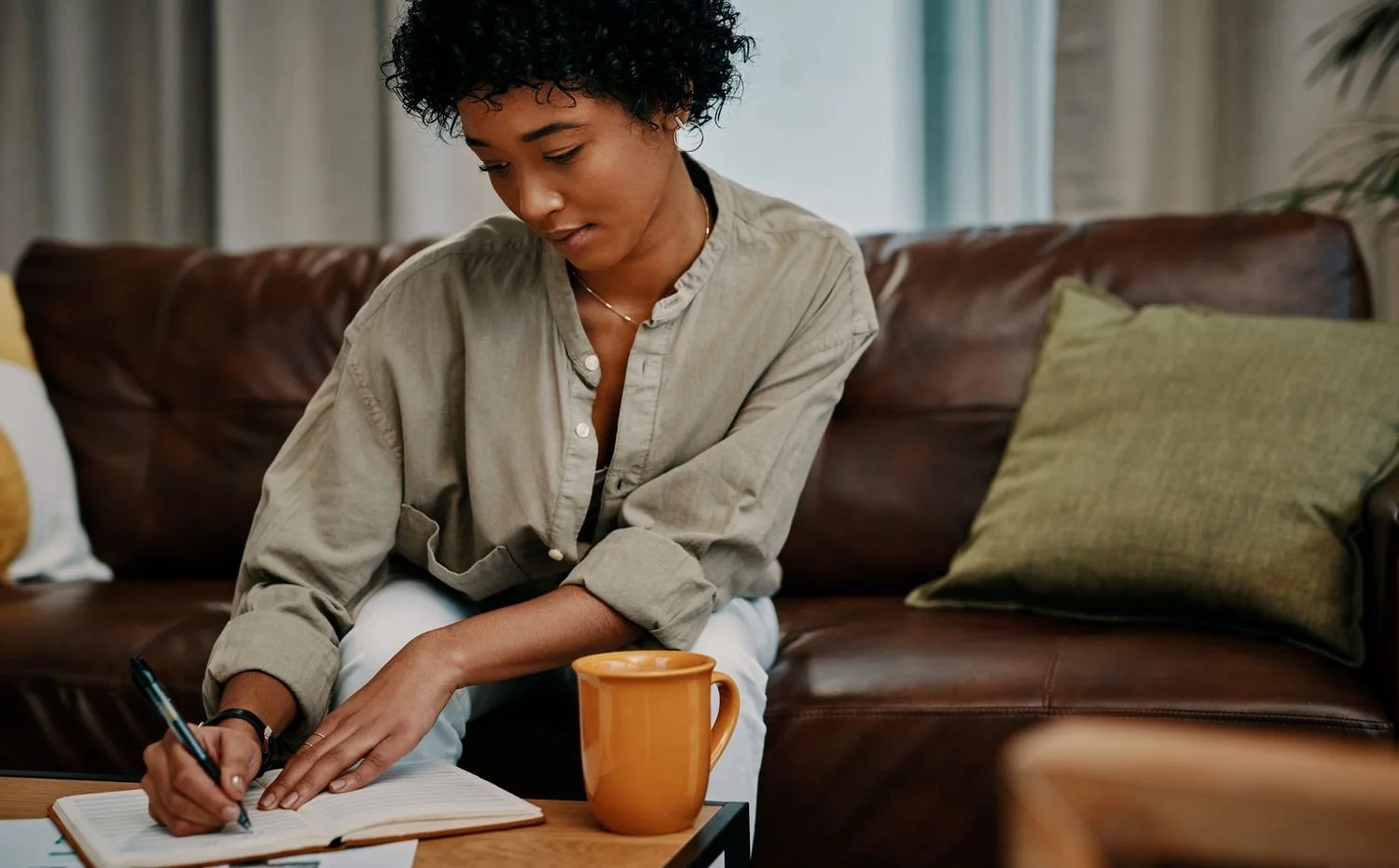 A woman sitting on a brown leather sofa, writing in a notebook with a pen, next to an orange coffee mug.
