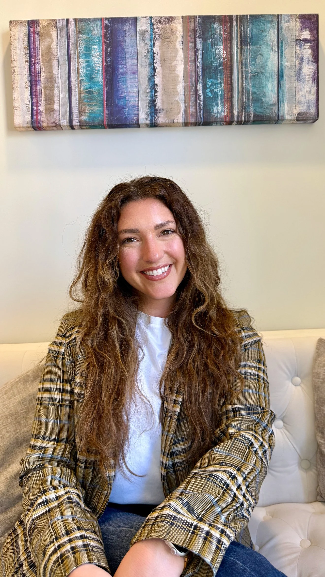 A woman with long curly brown hair smiling while sitting on a beige sofa, wearing a white t-shirt and a brown plaid blazer, with abstract artwork hanging on the wall behind her.