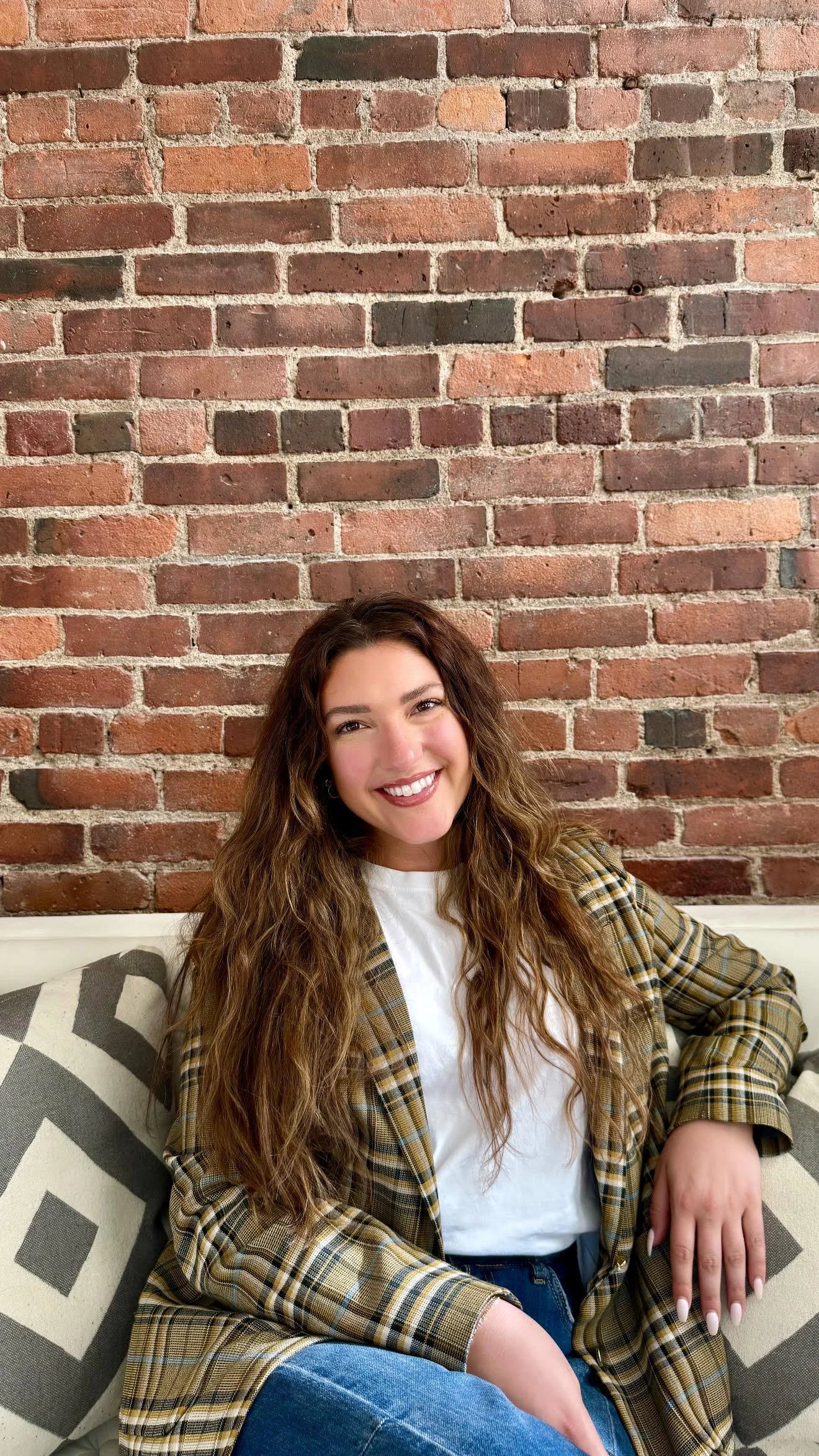 A woman with long, wavy brown hair sitting on a sofa with geometric patterned pillows, smiling, in front of a red brick wall.