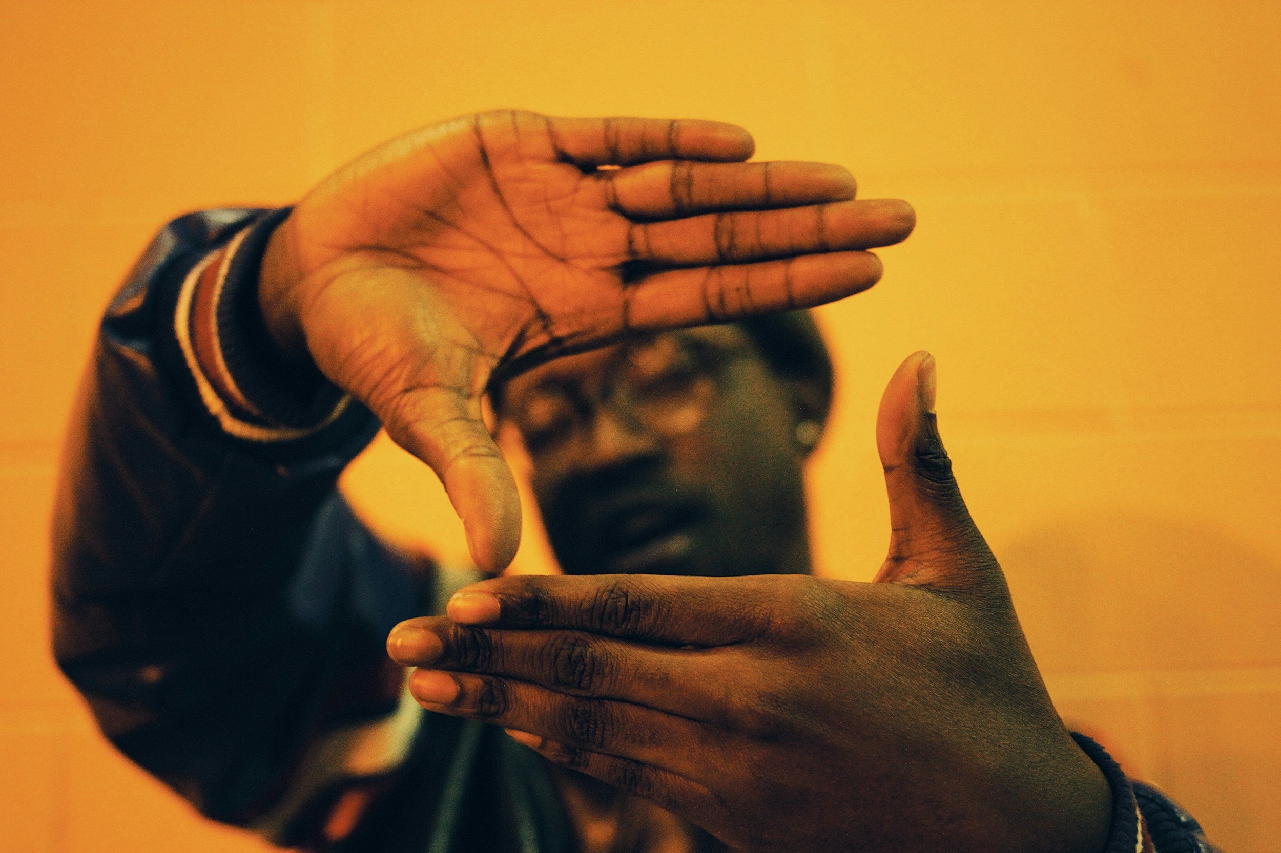 A person with dark skin, wearing glasses and a dark jacket with orange and white striped cuffs, is making a framing gesture with their hands against a yellow-orange background.