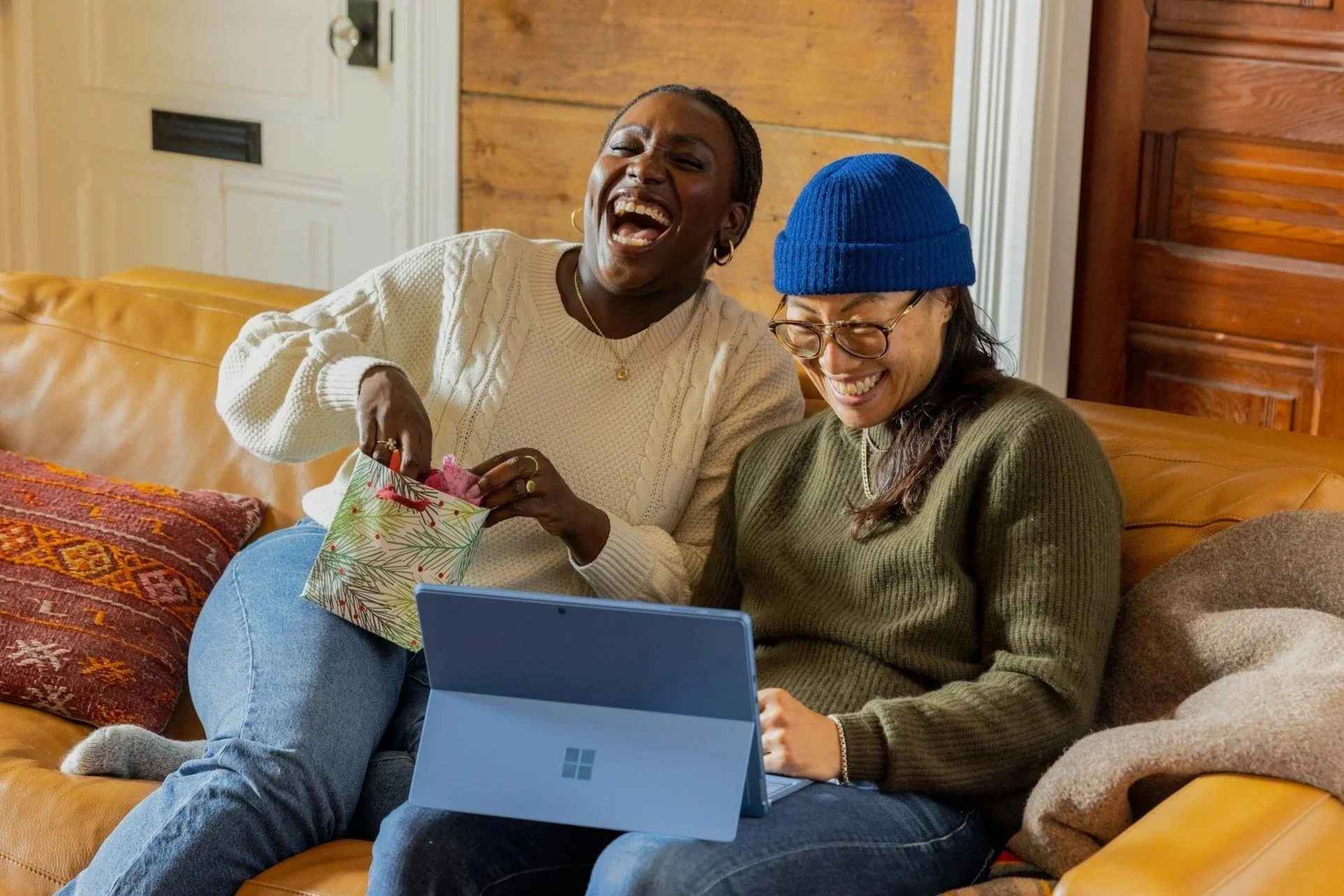 Two women sitting on a tan leather couch, smiling and laughing while looking at a tablet device. One woman is wearing a white sweater and the other a green sweater with a blue beanie, holding a gift bag.