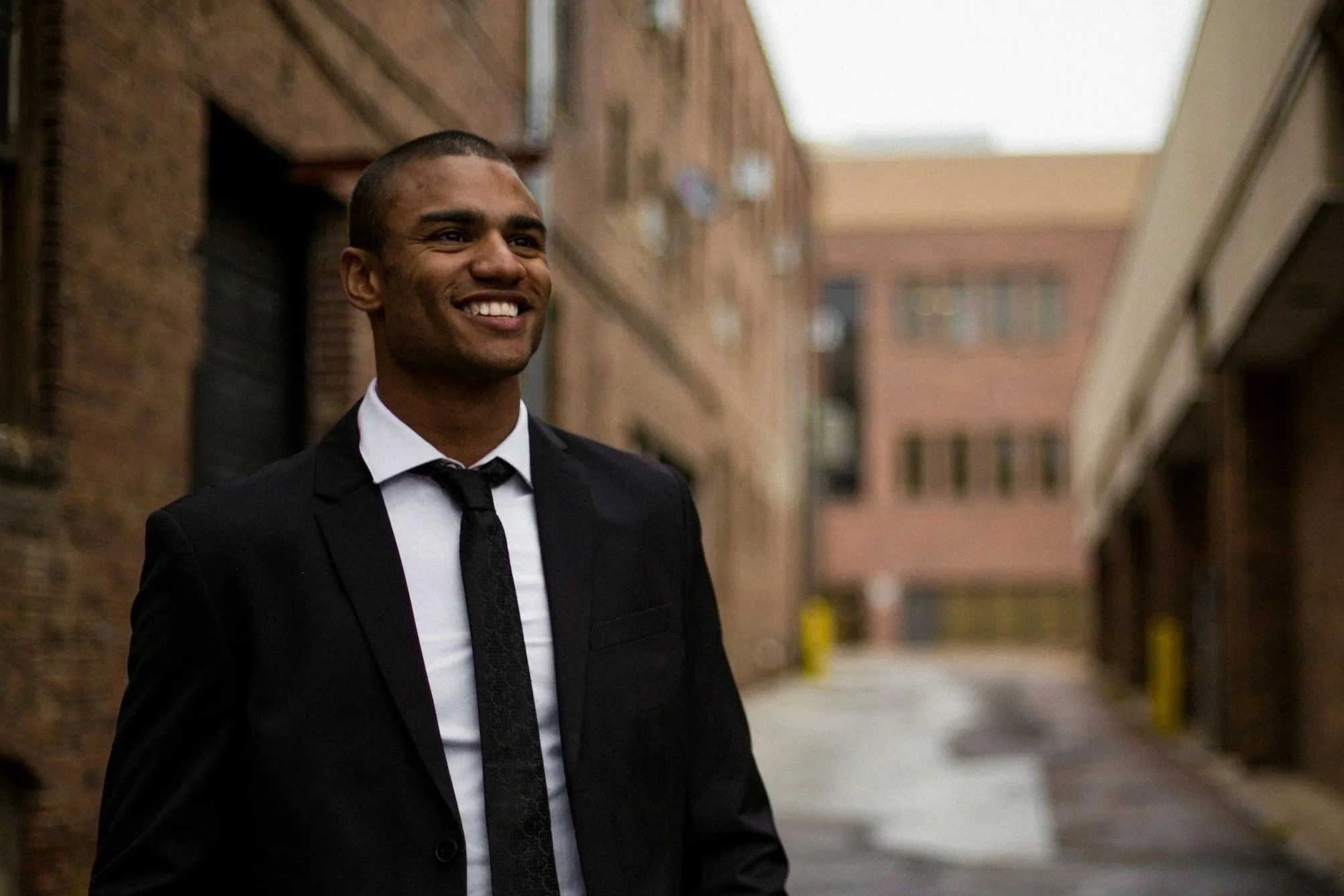 A smiling man in a black suit, white shirt, and black tie standing in an alleyway between brick buildings.