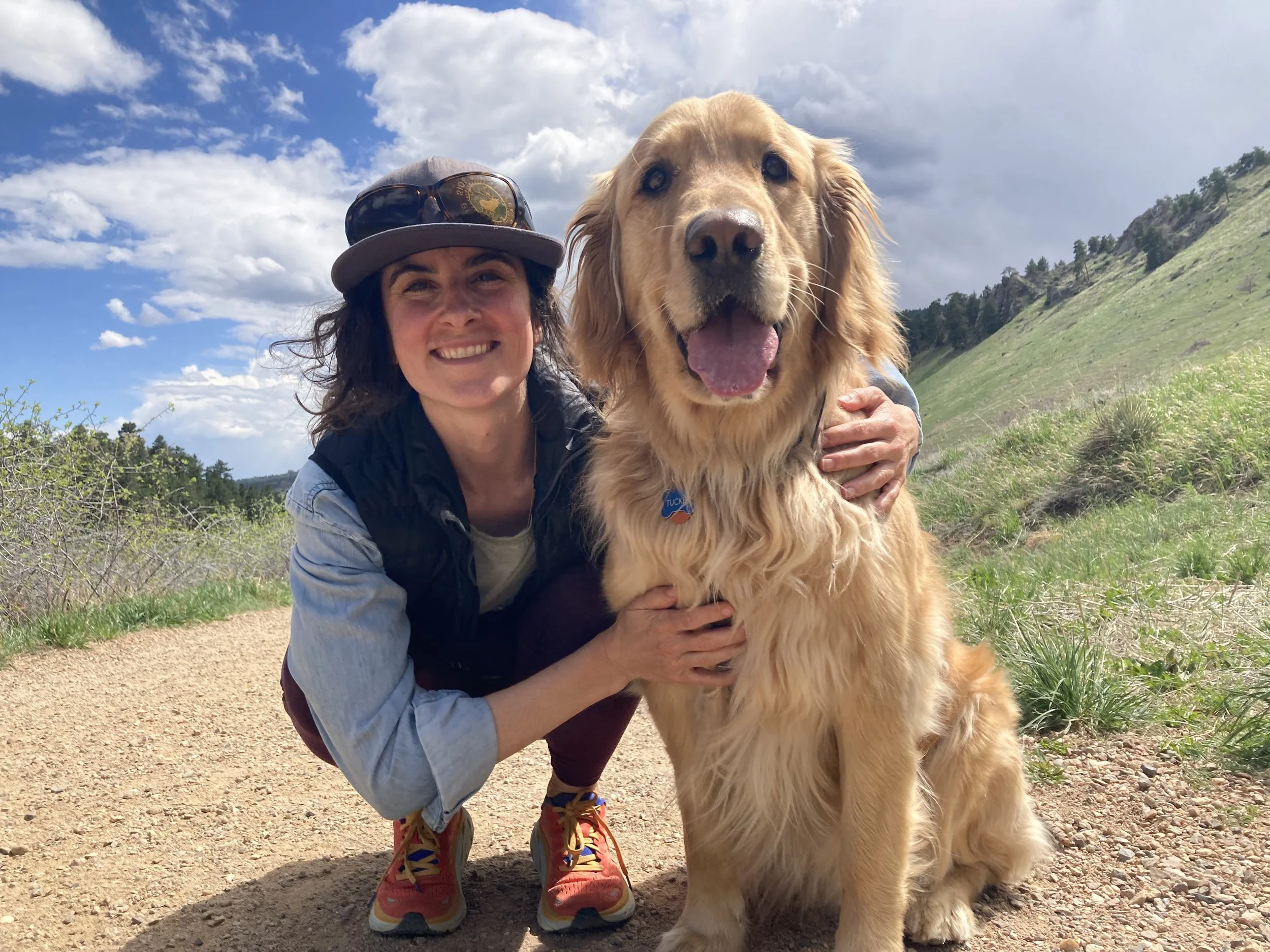 A woman squatting on a dirt trail outdoors, smiling, holding a large Golden Retriever dog with a blue tag, in a scenic hilly landscape with green grass, shrubs, and a partly cloudy sky.