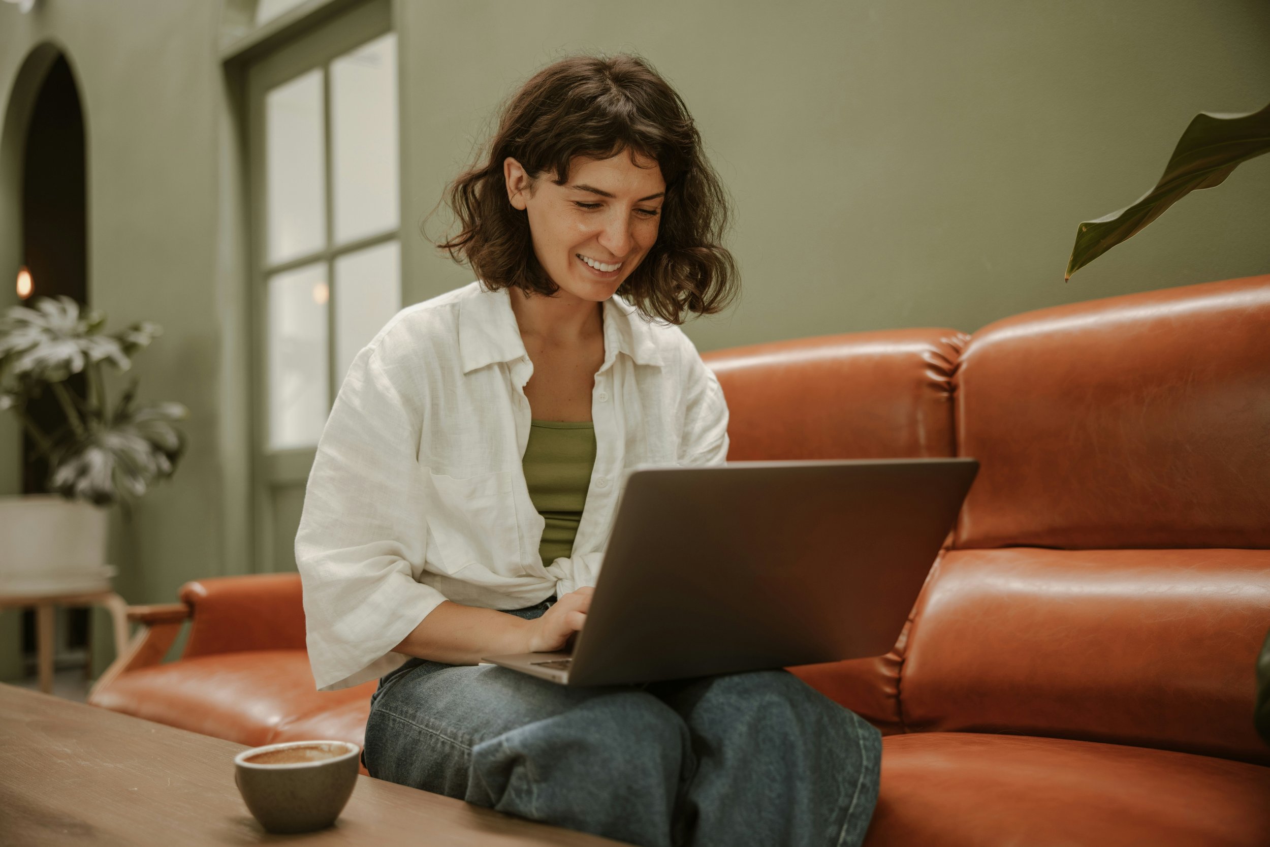 A woman with short brown hair, wearing a white open-collared shirt and jeans, sitting on an orange leather couch and smiling while looking at her laptop during a telehealth session for anxiety therapy.