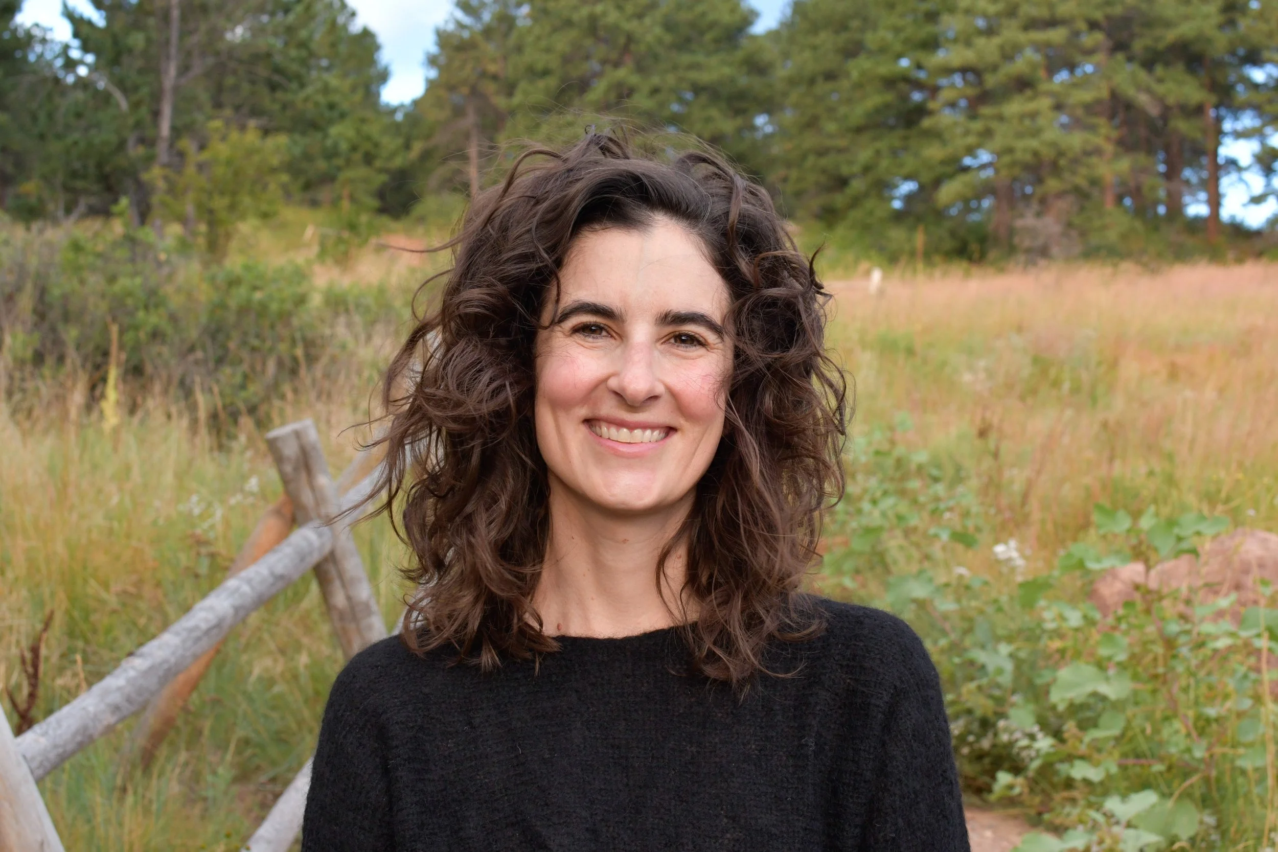 A woman with curly brown hair smiling outdoors in a grassy field.