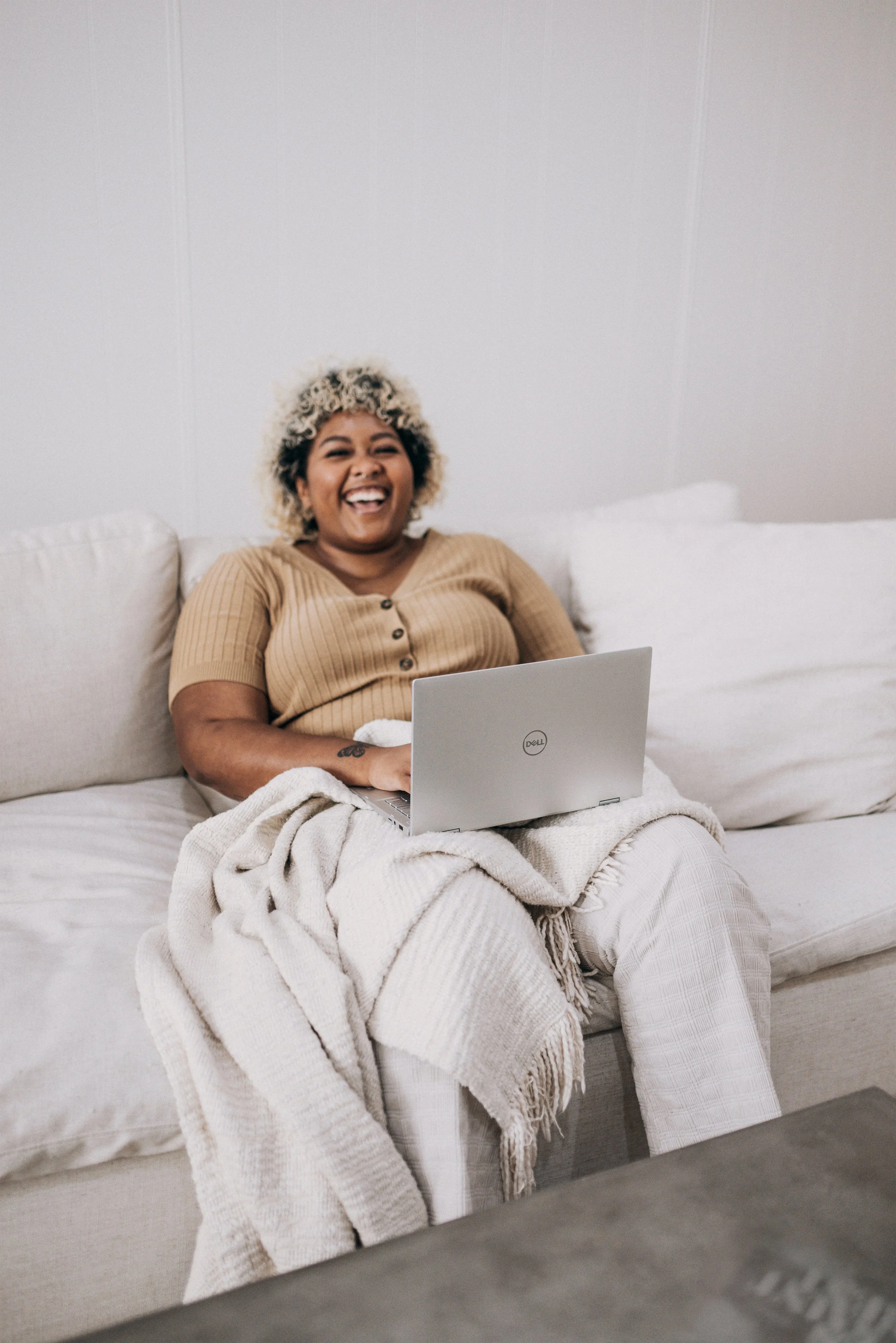 A woman with curly blonde hair and a big smile sitting on a beige couch with a laptop on her lap in a cozy room, wearing a tan shirt, with a beige blanket covering her legs.