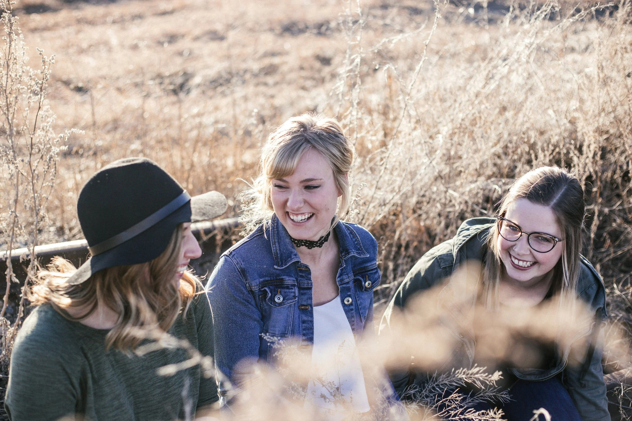 Three women sit smiling and laughing in a field of long golden grass talking about the benefits of trauma therapy in Colorado.