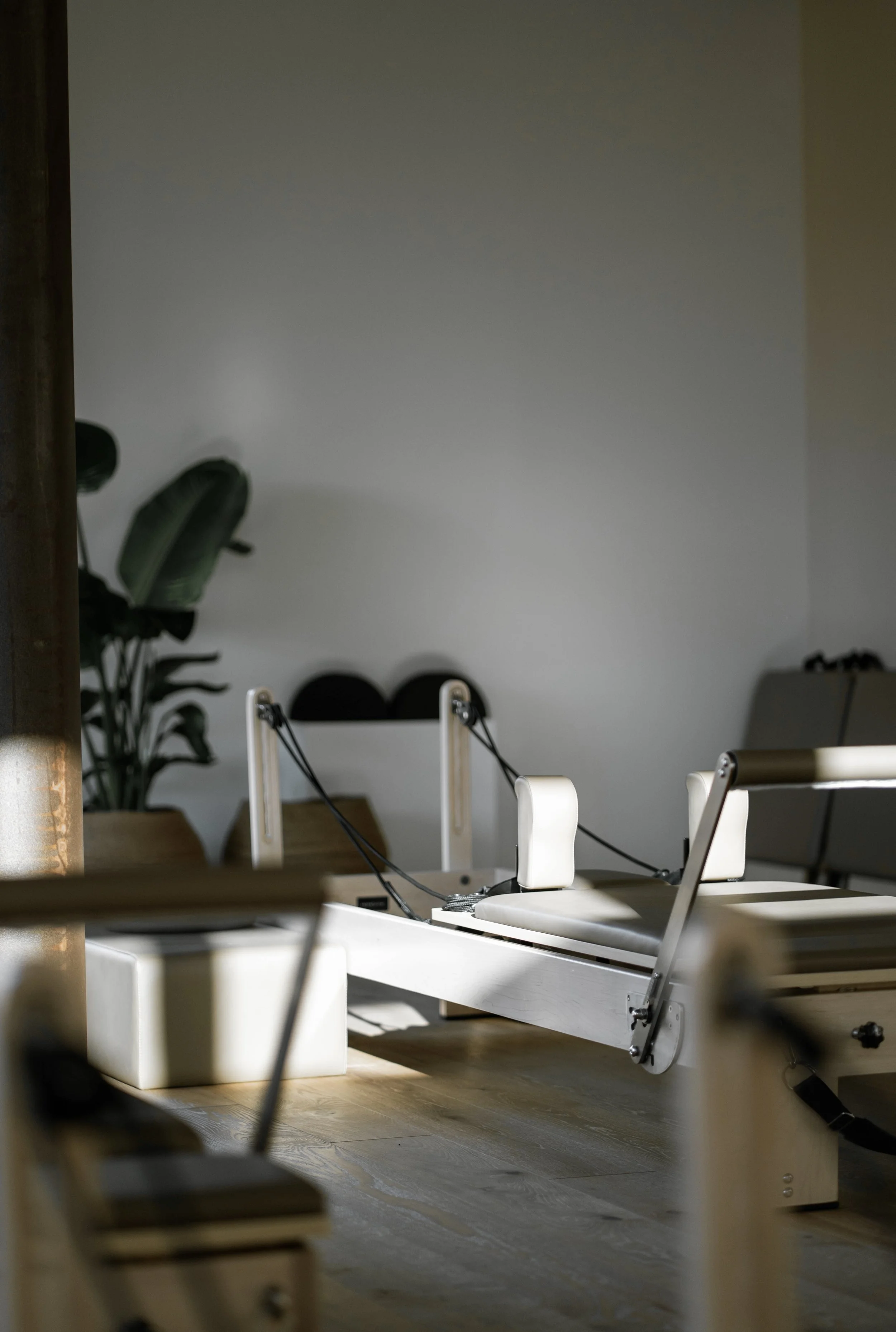 Ein liegendes Rehabilitationstraining-Gerät in einem Raum mit Holzboden, Licht und Schatten. Im Hintergrund eine Pflanze sowie Stühle und ein Sofa.