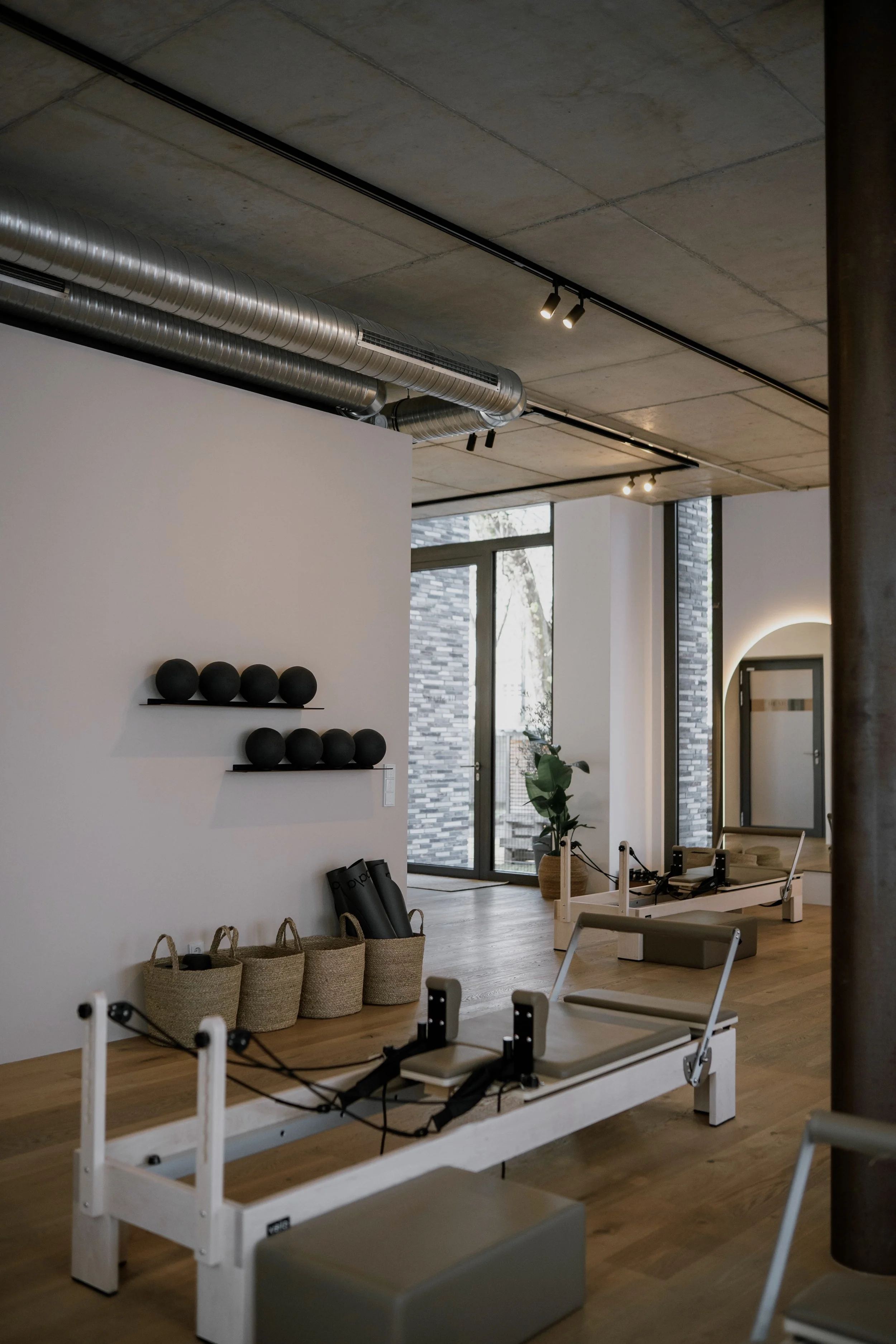 Innenraum eines Fitnessstudios mit Pilates- und Trainingsgeräten, schwarzen Bällen an der Wand, Korbgläsern mit Handtüchern und Pilates-Equipment, viel natürlichem Licht durch große Fenster.