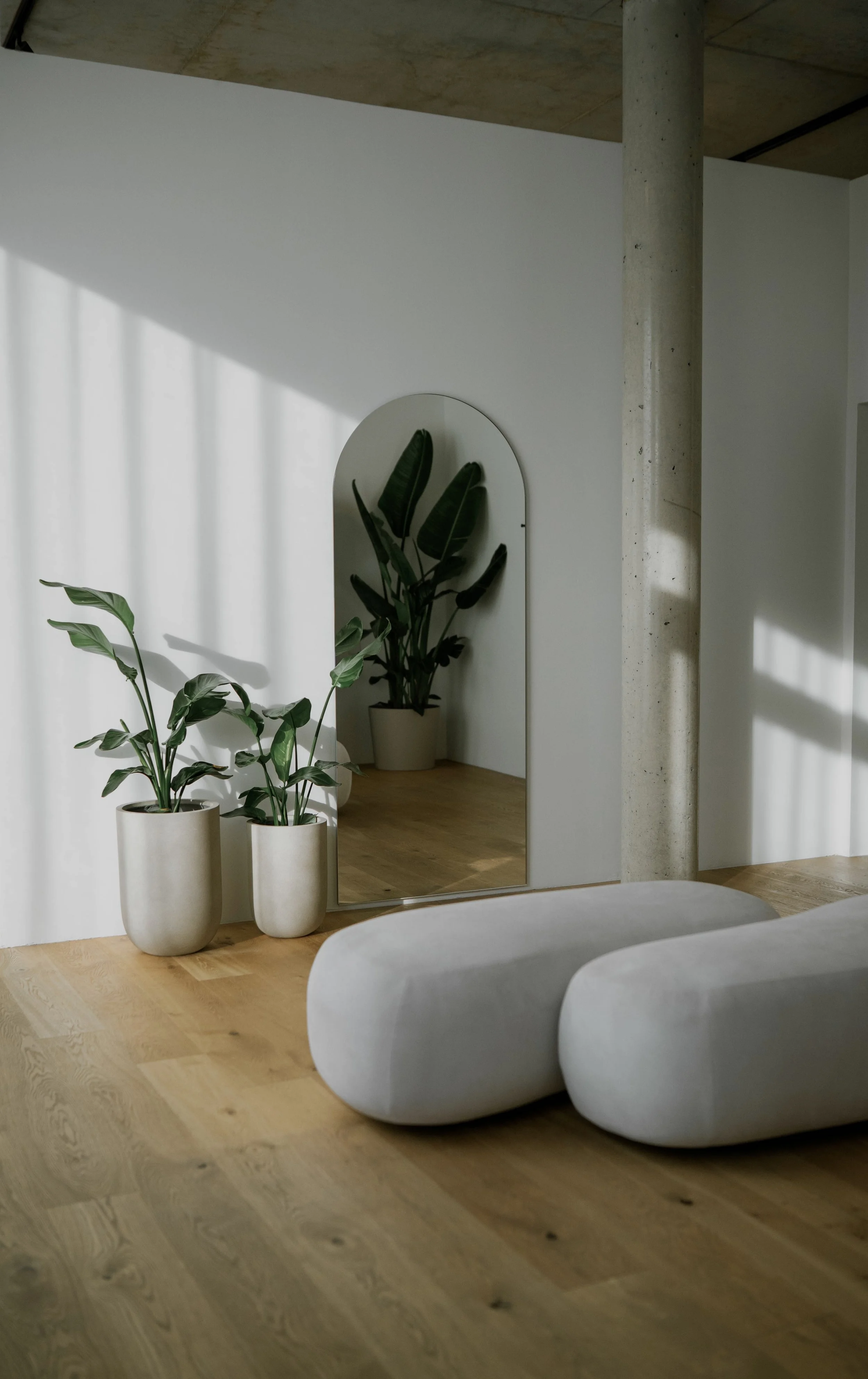 Minimalistischer Raum mit Holzboden, zwei weiße, organisch geformte Sitzmöbel, ein dekorativer Spiegel mit Bananenpflanzen in weißen Töpfen, natürliches Licht mit Schattenmustern an der weißen Wand.