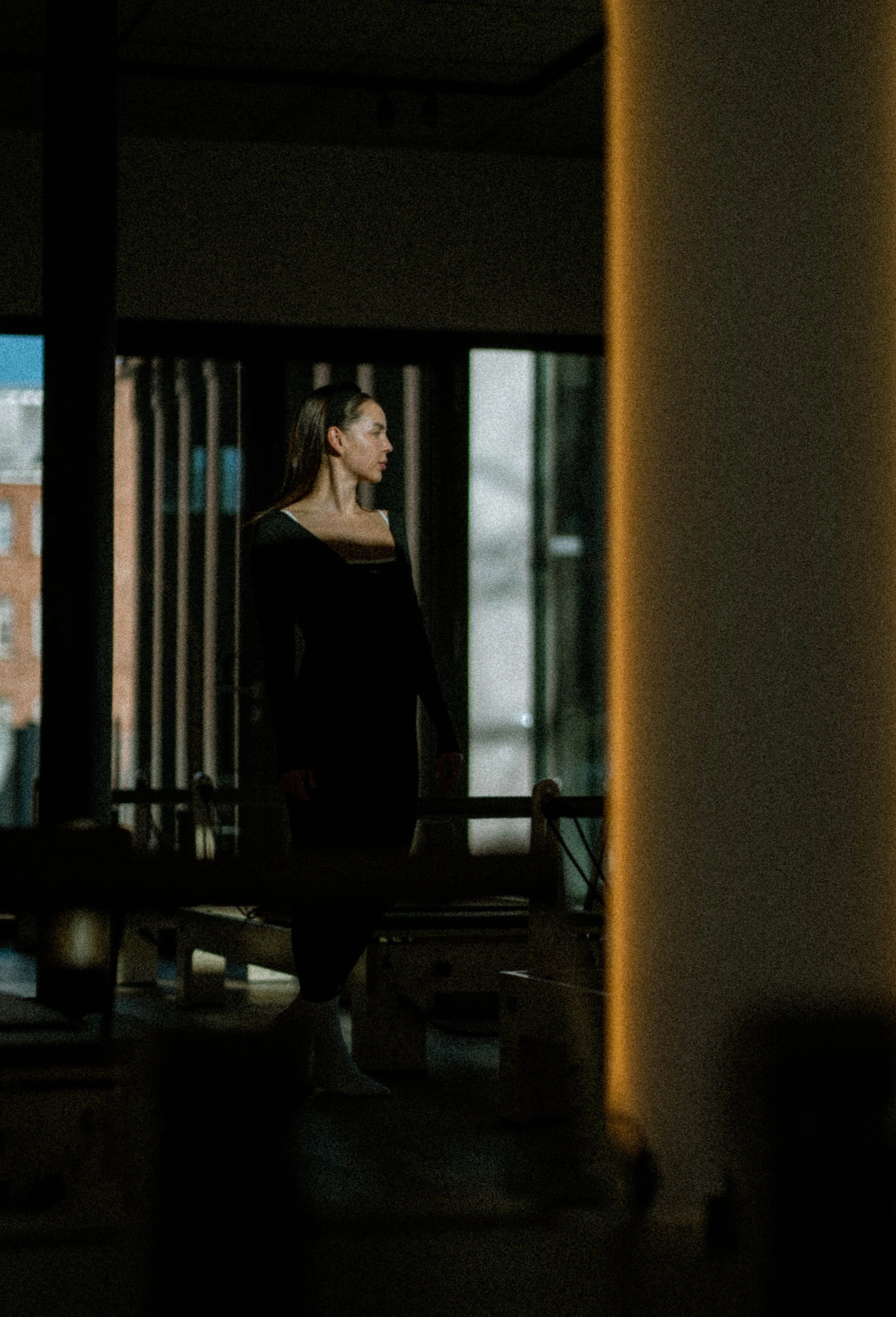 Eine Frau in schwarzer Kleidung steht in einem dunklen Raum neben einem Fenster. Es ist dunkel draußen, sichtbar durch das Fenster.