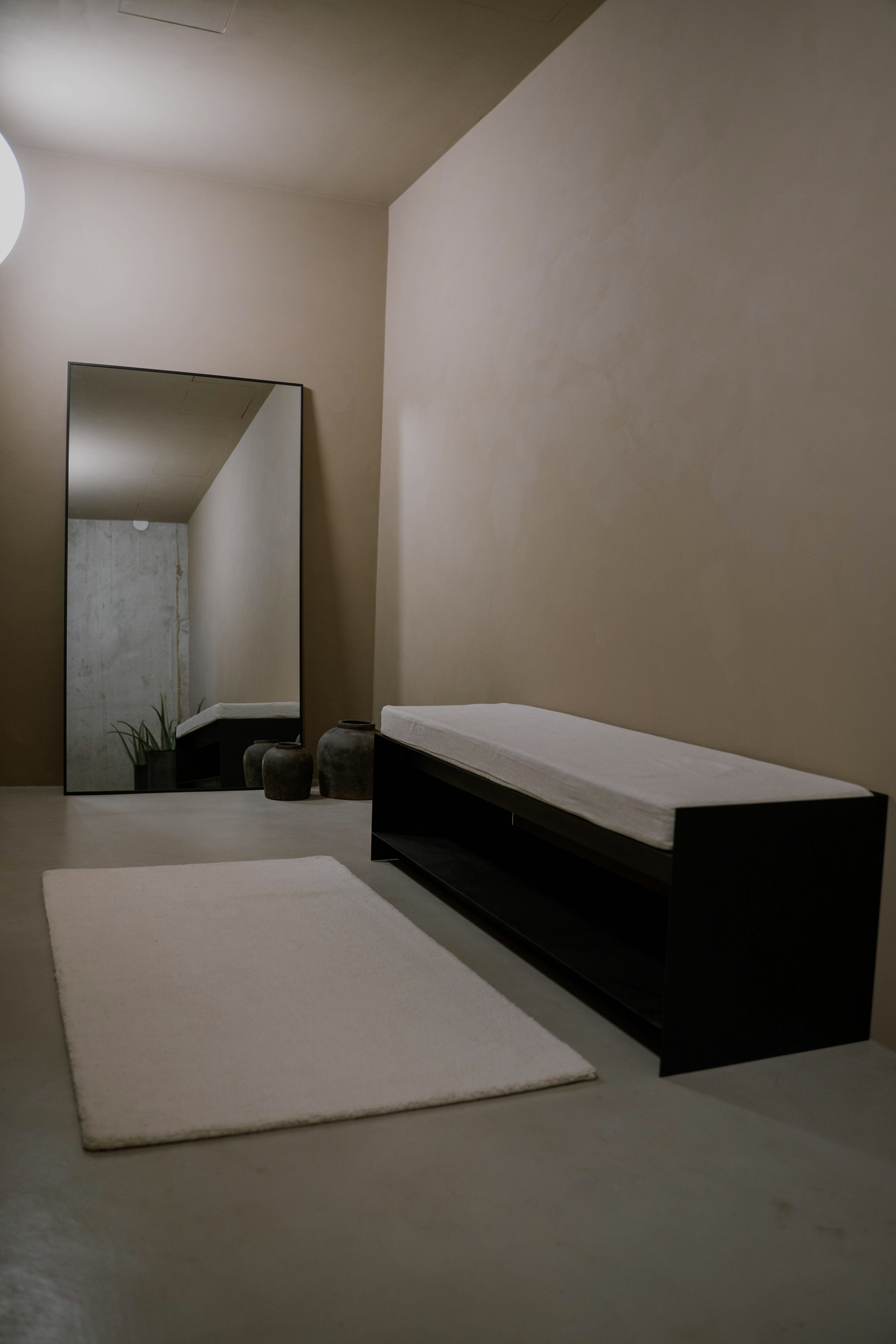 Minimalistischer Eingangsbereich mit großer Spiegelreflexion, schwarzem Sitzbank mit weißen Kissen, weißen Teppich, drei Keramikvasen, einer Pflanze und neutralen Farbtönen.