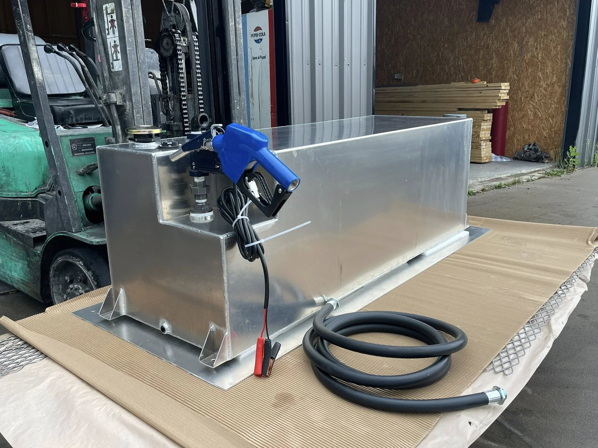 A large metal container with hoses and electrical equipment on top, placed on a sheet of cardboard in an industrial or workshop setting.